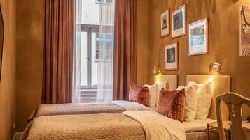 Ein Hotelzimmer mit zwei Betten, einer Chandelier, einem großen Fenster mit Vorhängen und gerahmten Bildern an der Wand.