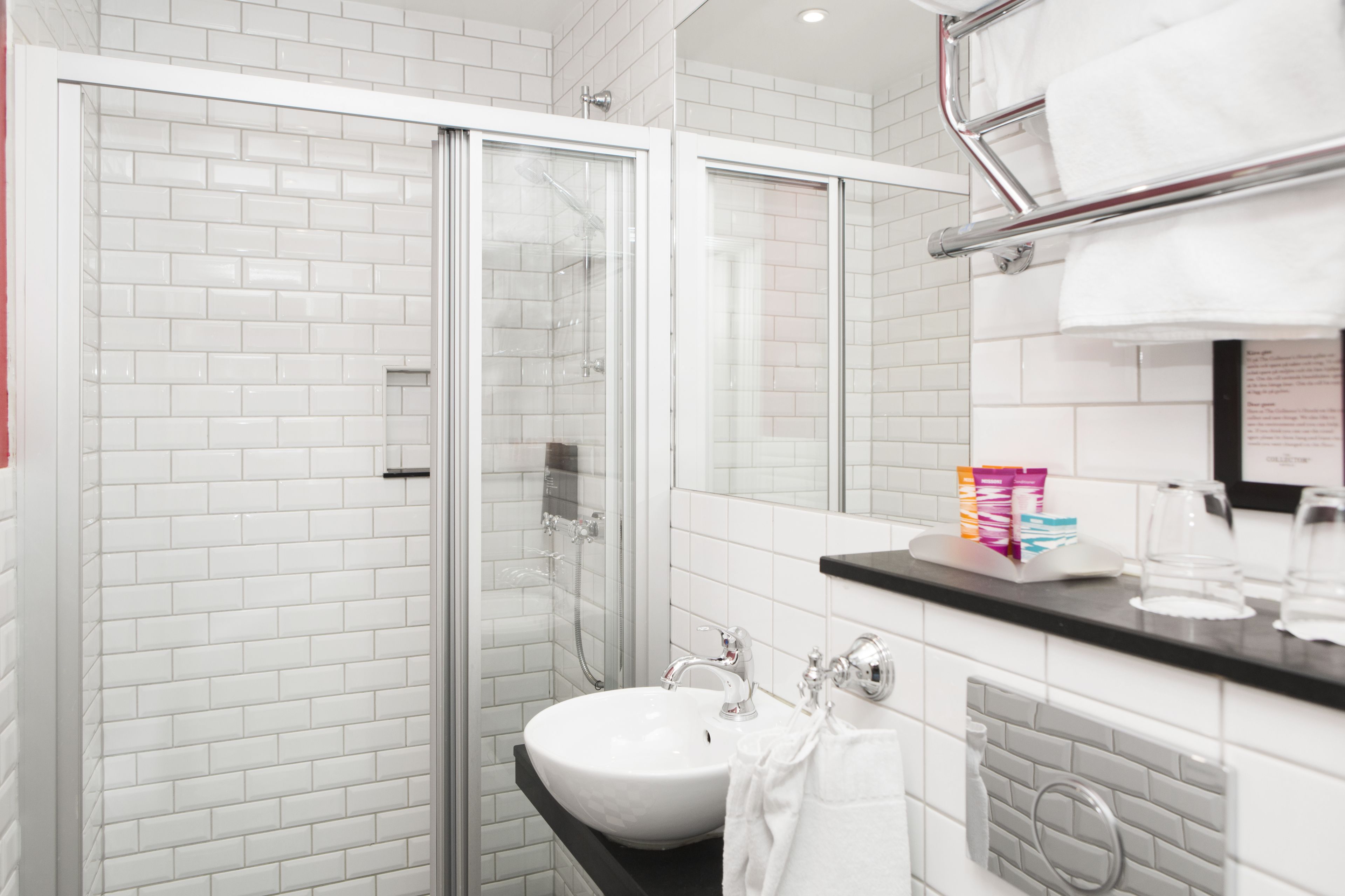 Een moderne badkamer met witte metrotegels, een glazen douchecabine, een witte wastafel en een zwart aanrechtblad met toiletartikelen.