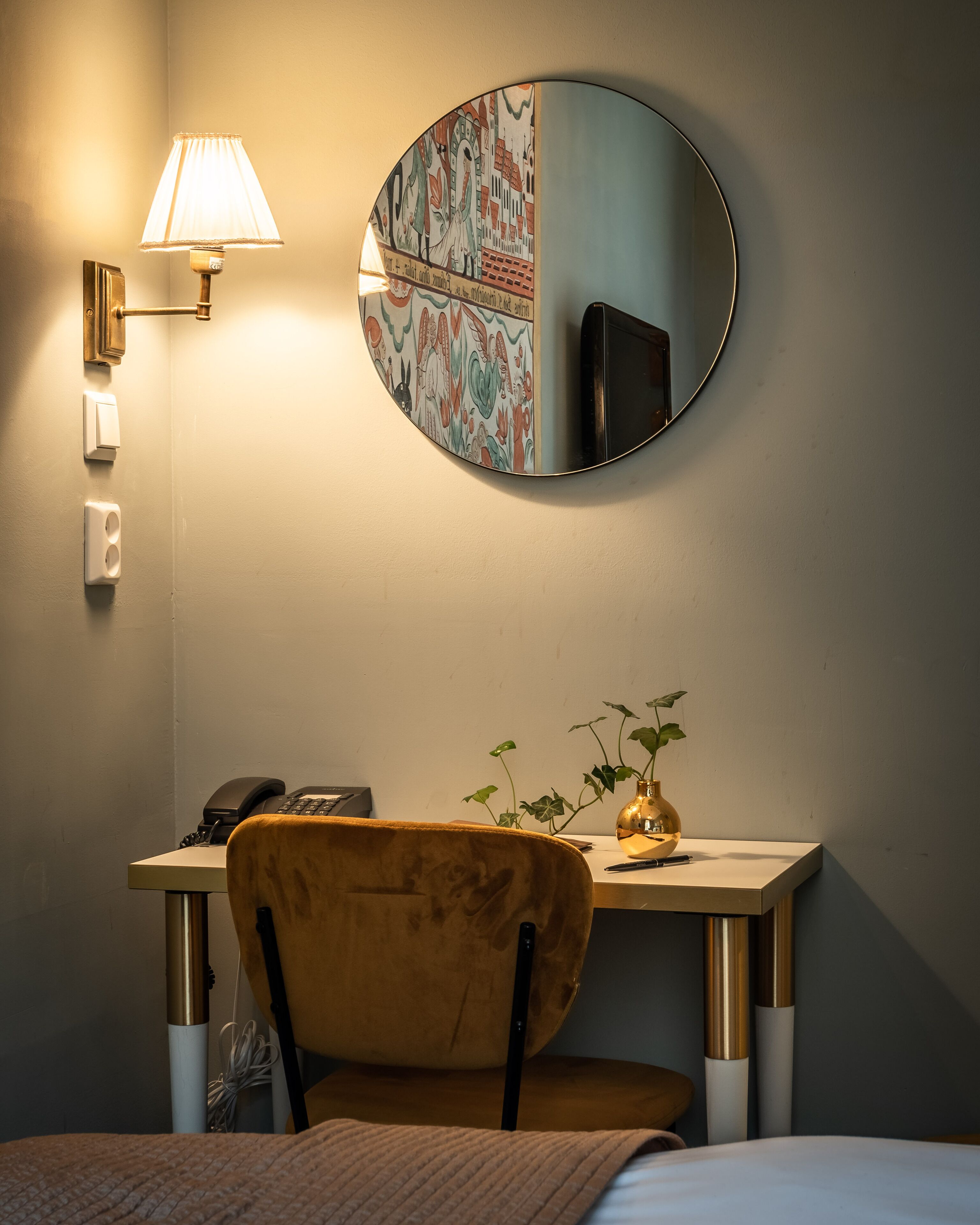 A hotel room with a round mirror, a desk with a telephone, a chair, and a bedside table with a plant.