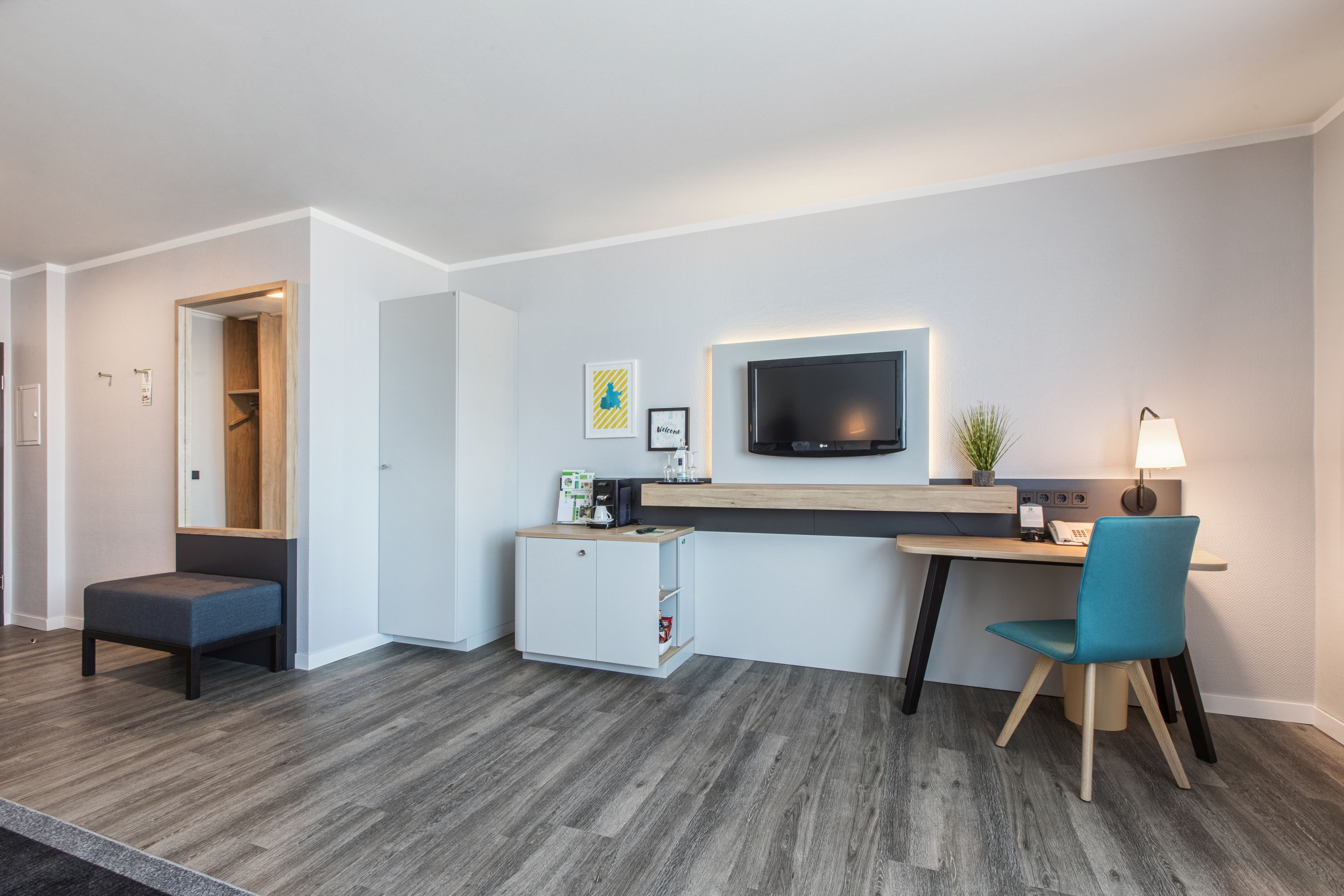 A modern hotel room with a kitchenette, a flat-screen TV mounted on the wall, a desk with a chair, and a small seating area.