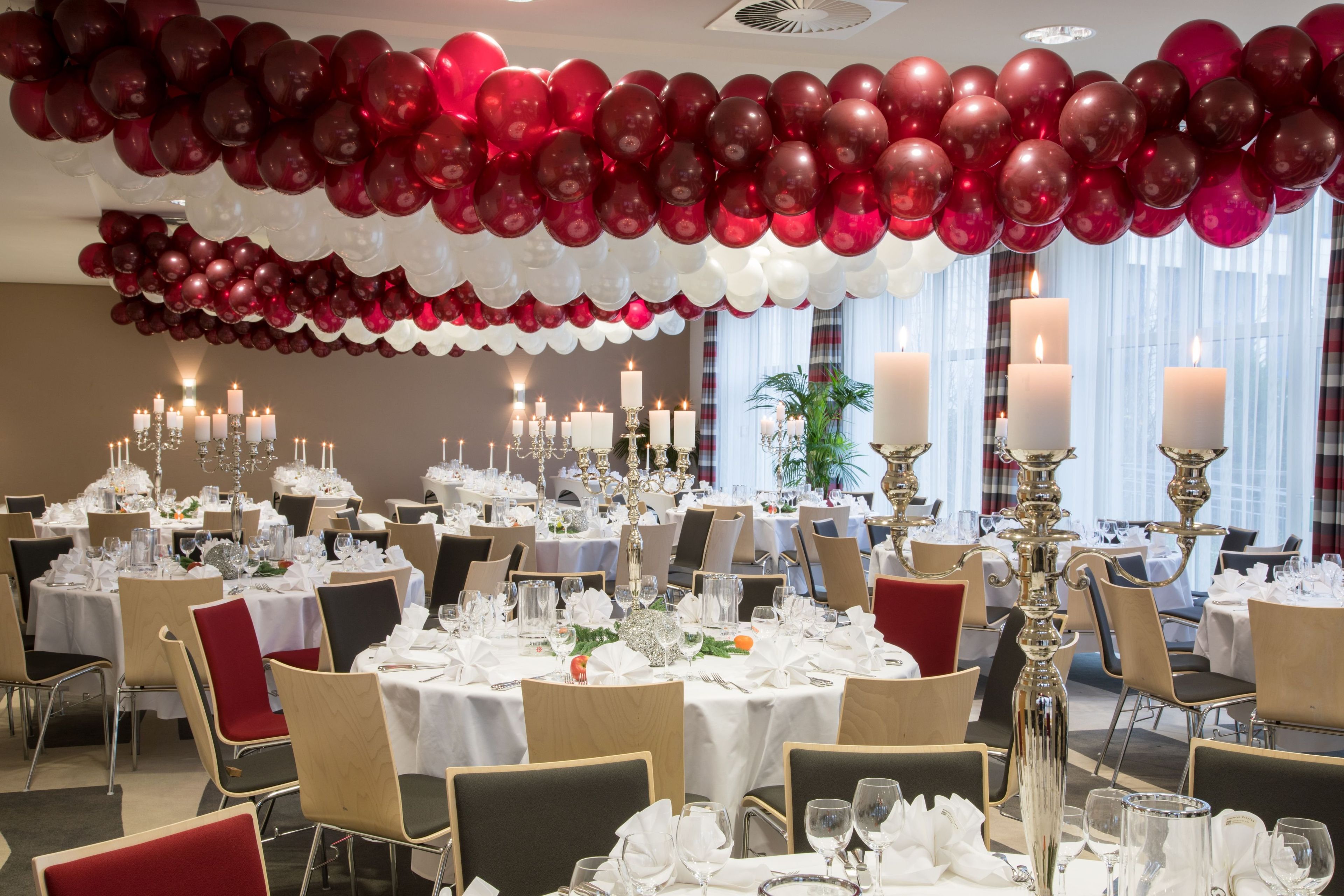 Un salón de banquetes con mesas redondas dispuestas para cenar, un techo decorado con globos rojos y blancos, y varios candelabros.
