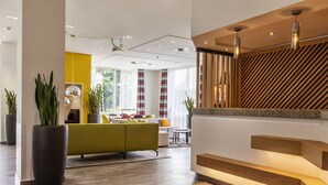 Un vestíbulo de hotel moderno con una escalera de madera, un sofá verde y grandes macetas con plantas.
