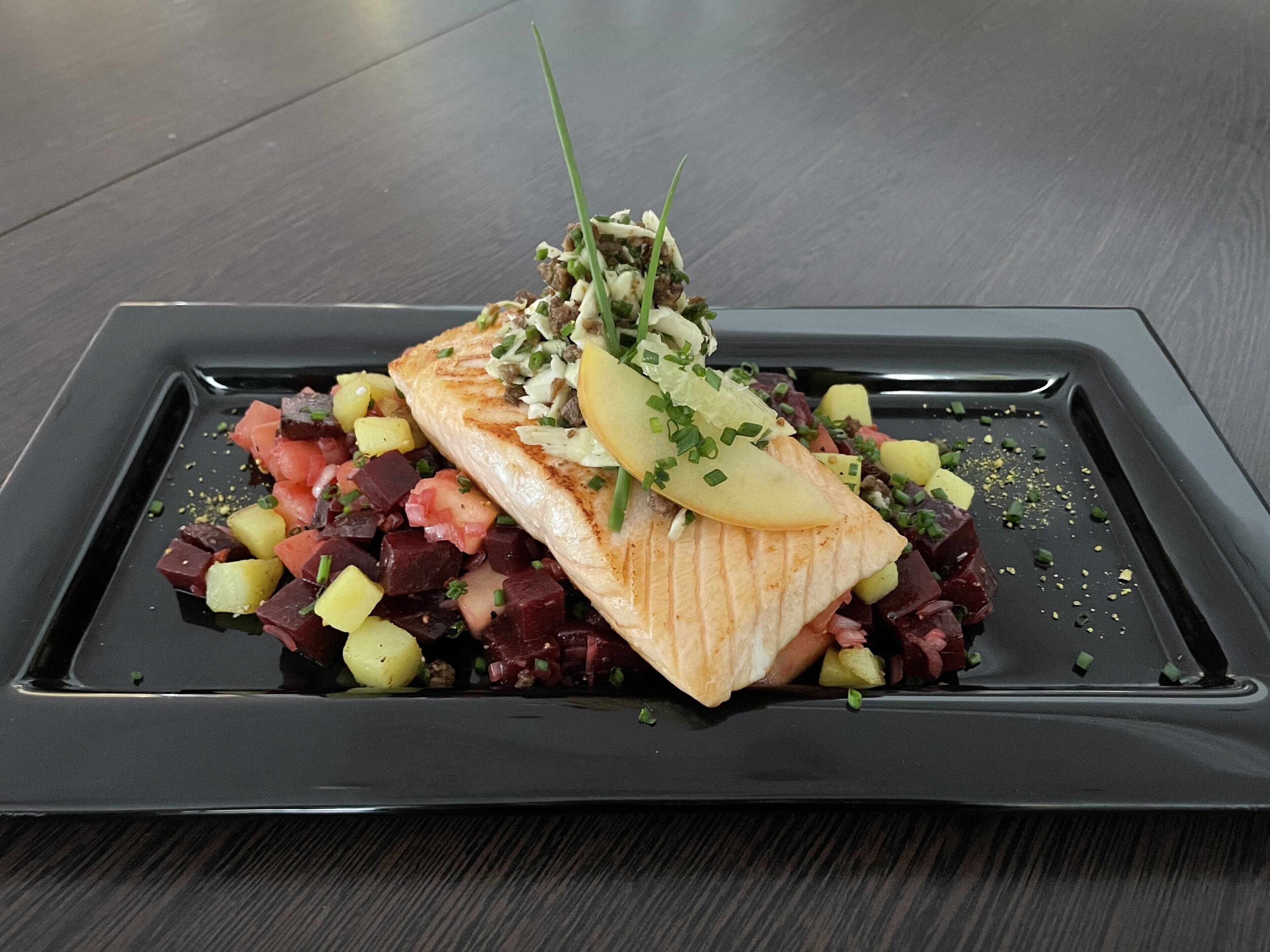 A rectangular plate with a piece of grilled fish, diced vegetables, and garnishes on a dark surface.