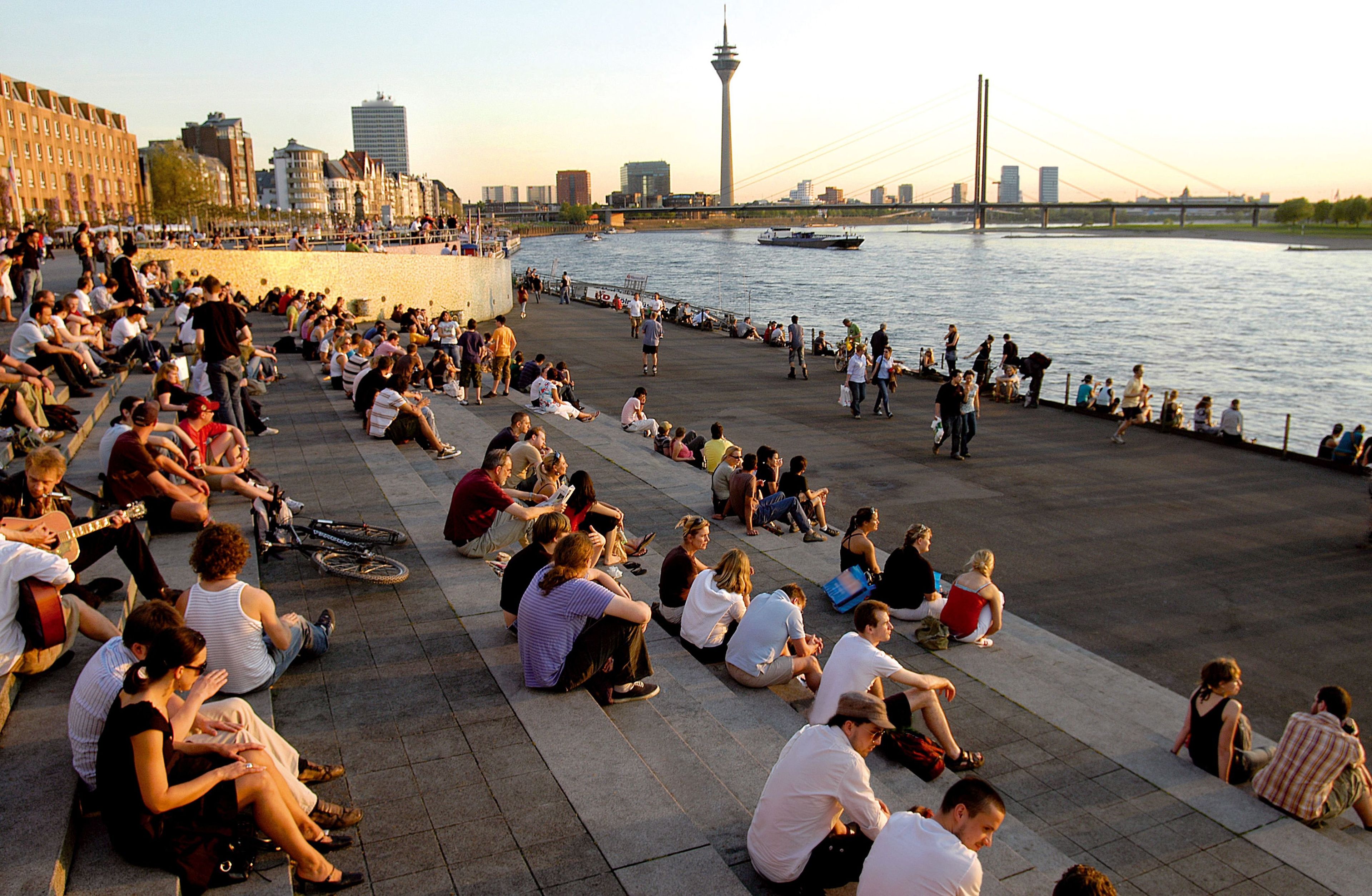 A riverside promenade with people sitting and walking, a bridge in the background, and a tower.