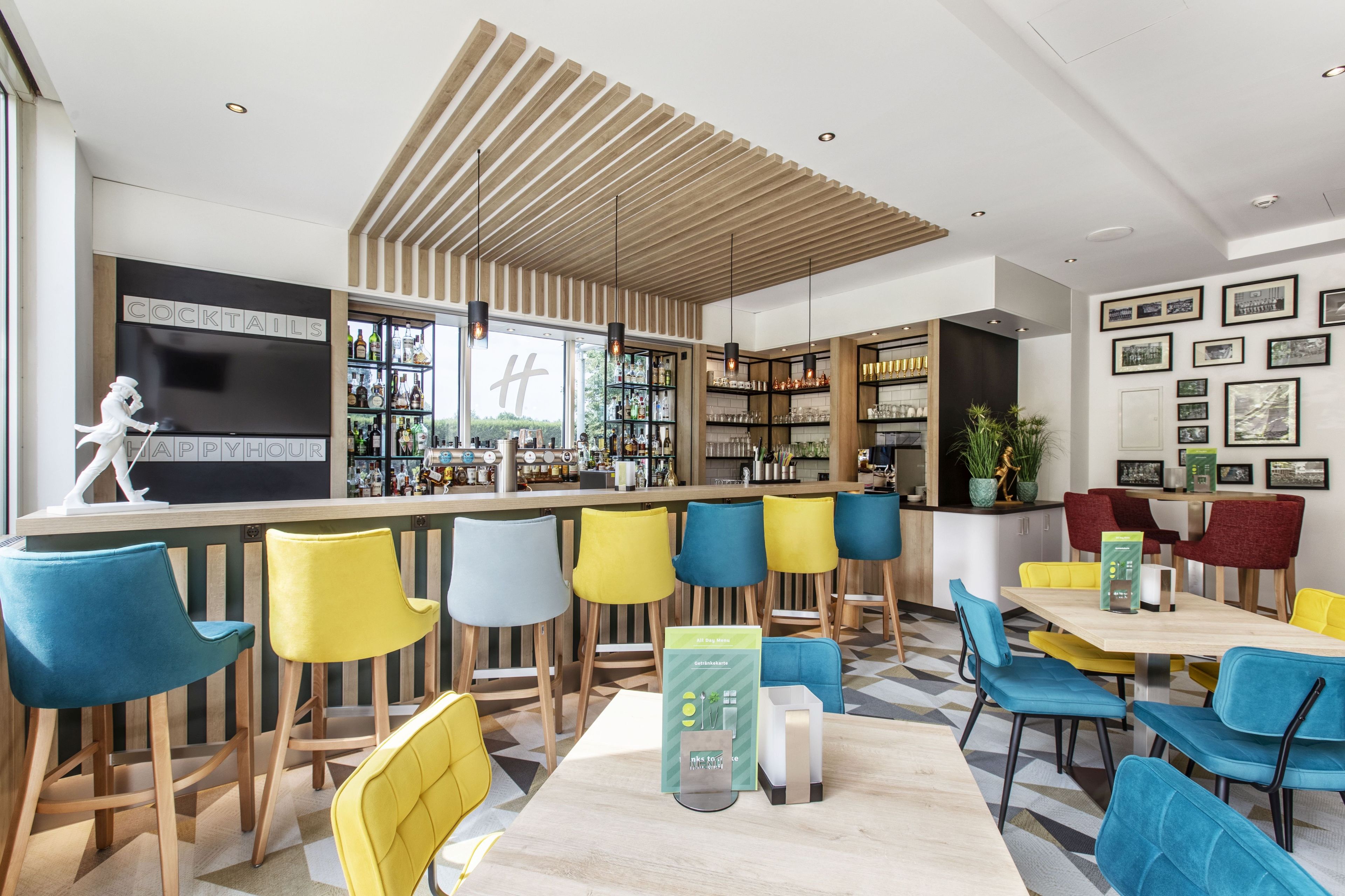 A modern bar area with wooden slats on the ceiling, a bar counter with a variety of bottles, and colorful bar stools.