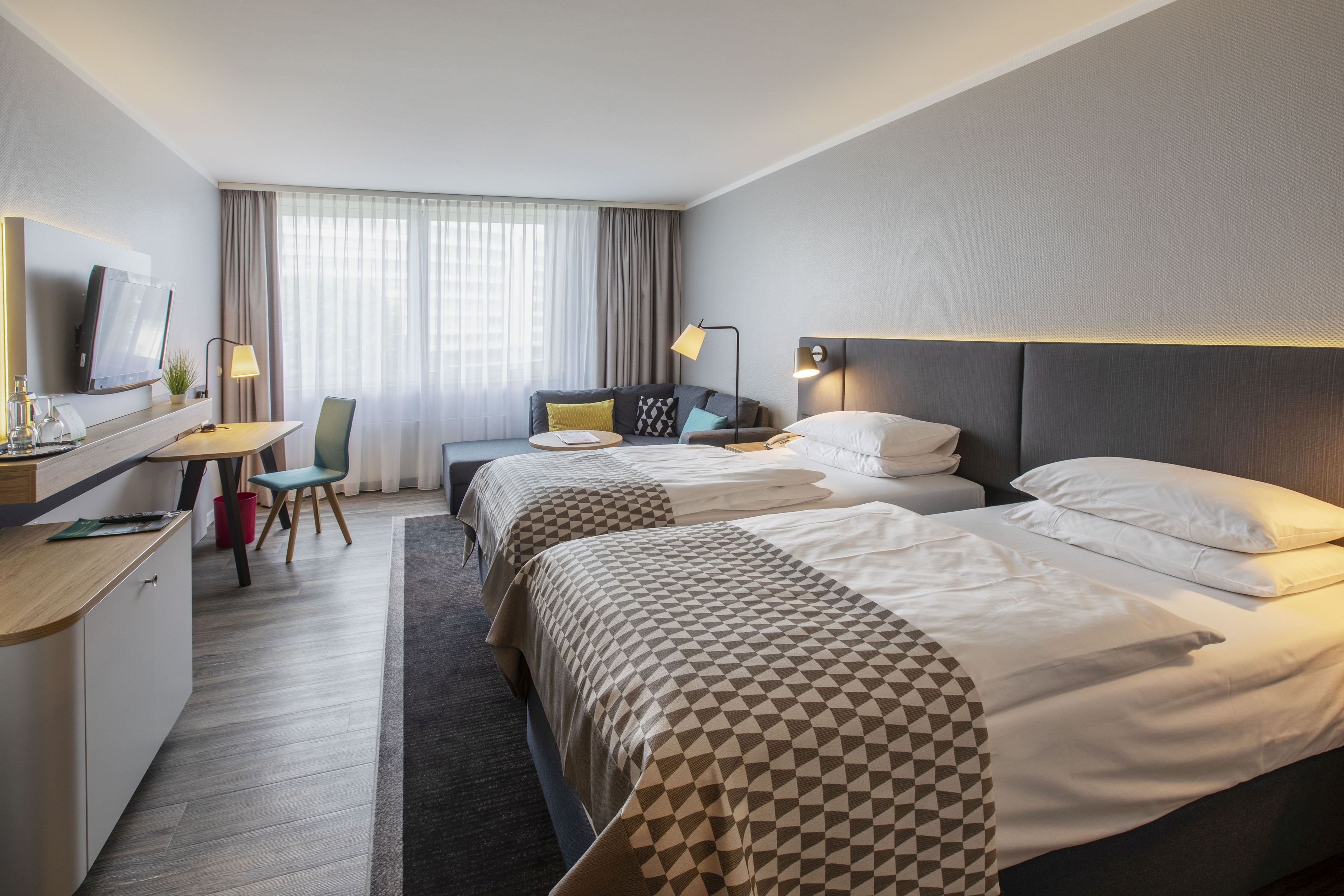 A hotel room with two beds, a desk, a chair, a TV, and a window with curtains.
