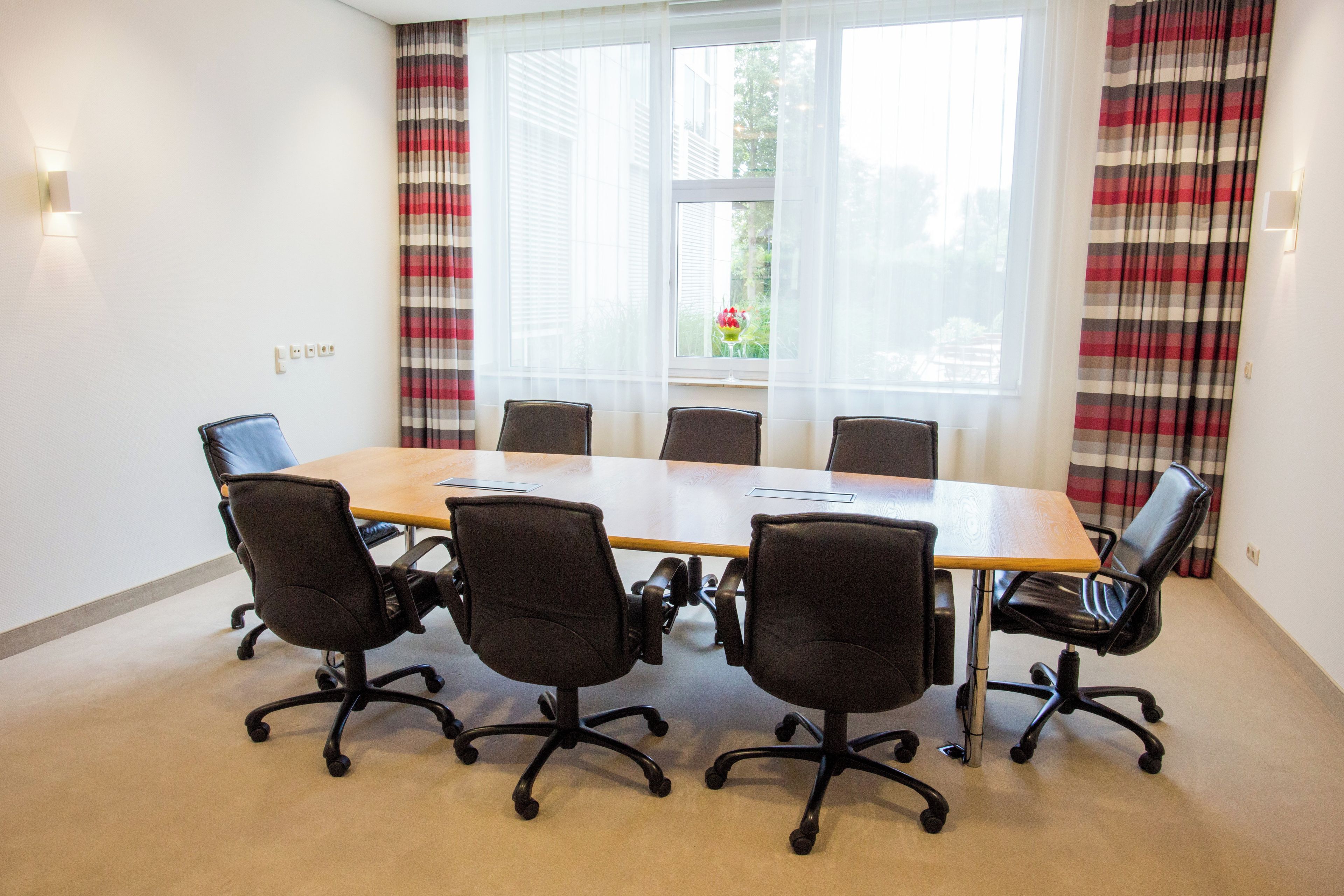 A conference room with a wooden table, six black office chairs, and a large window with curtains.