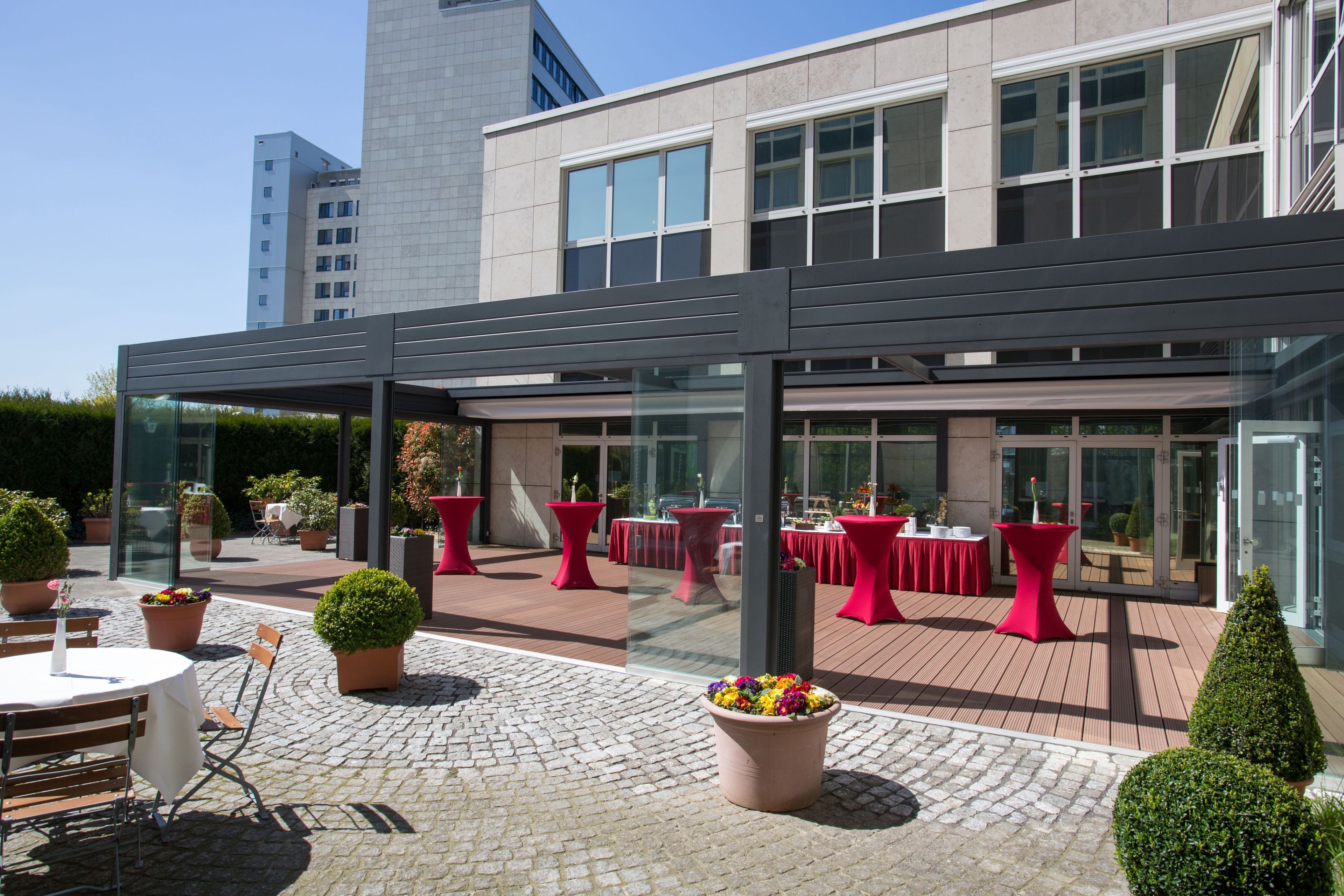 A modern outdoor patio area with tables and chairs, potted plants, and a glass enclosure.