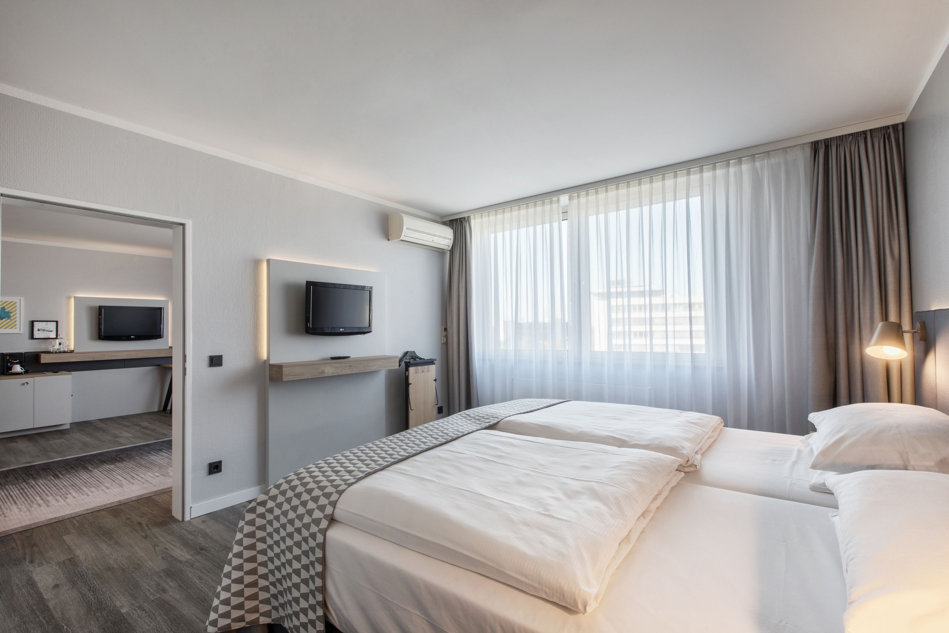 A hotel room with a bed, a desk with a TV, an air conditioning unit, and a window with curtains.