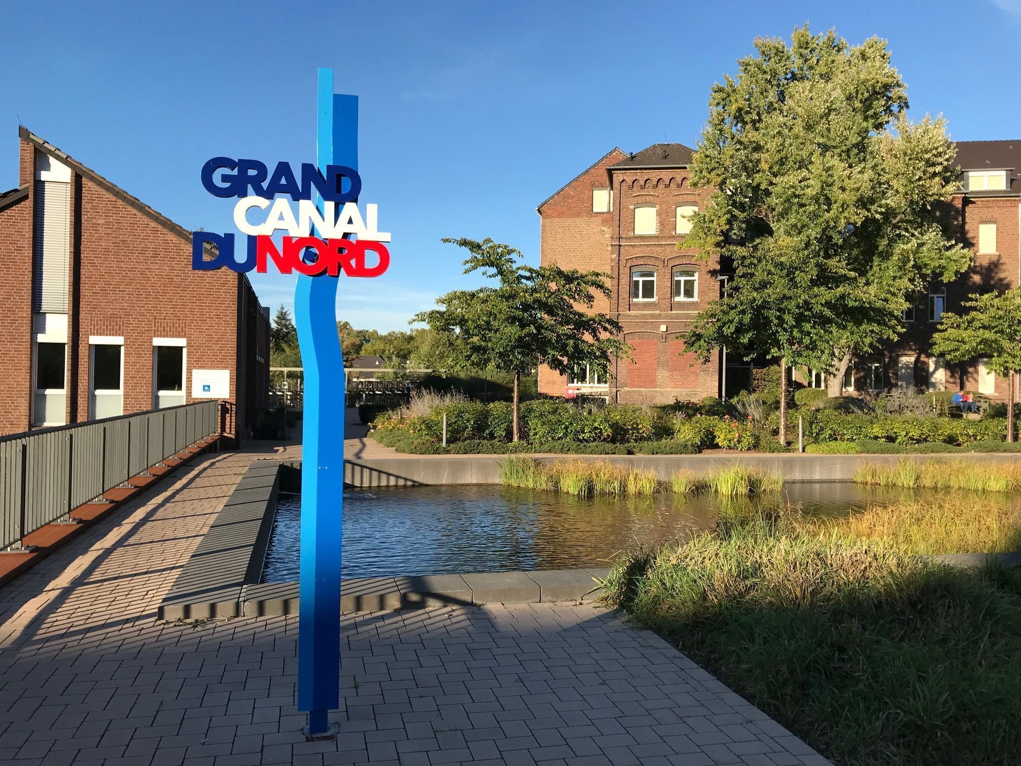 A sign for Grand Canal Bund, a canal-side walkway with a bridge, a water body, and buildings in the background.