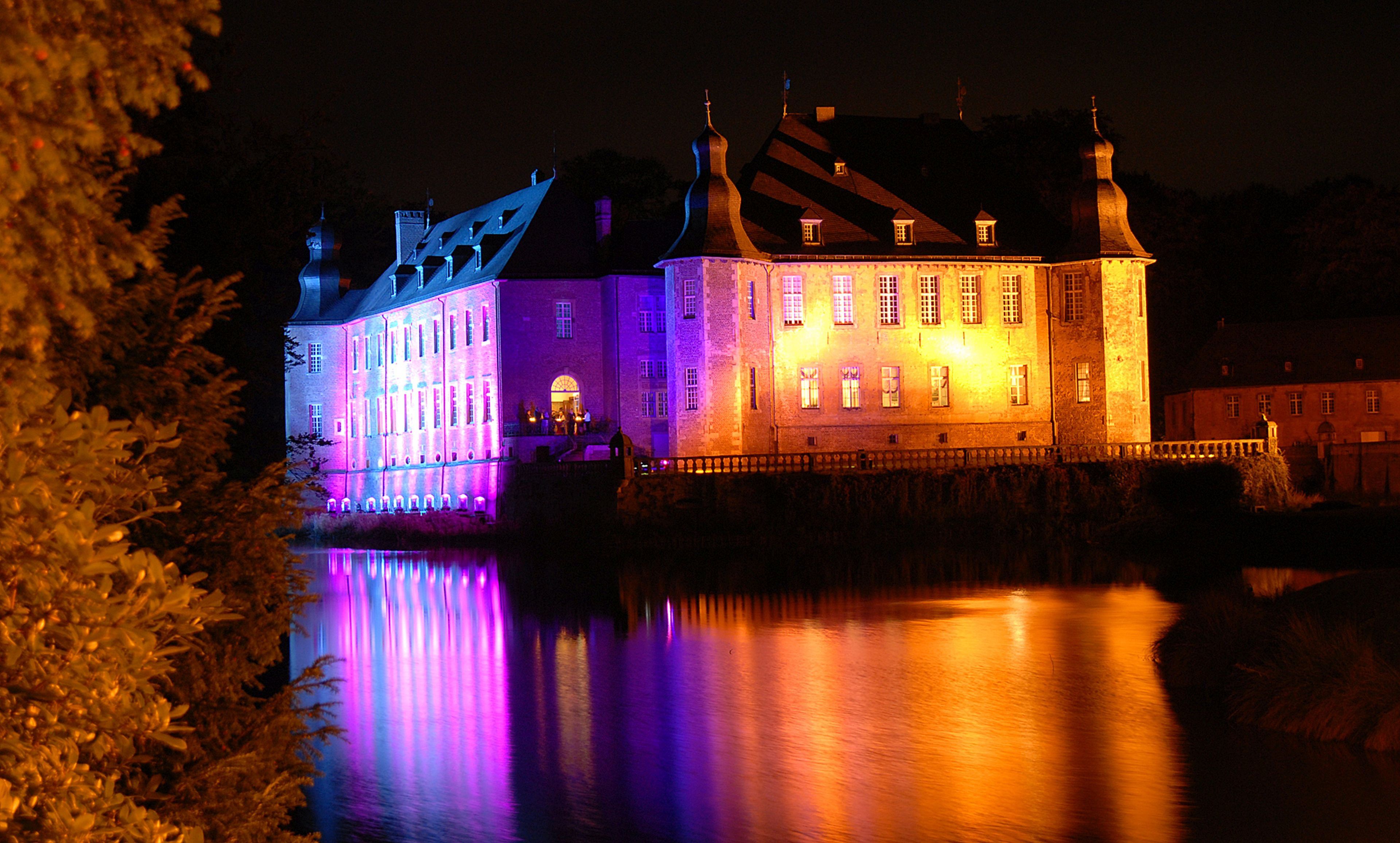 A historic building with multiple towers and a moat, illuminated by colorful lights.