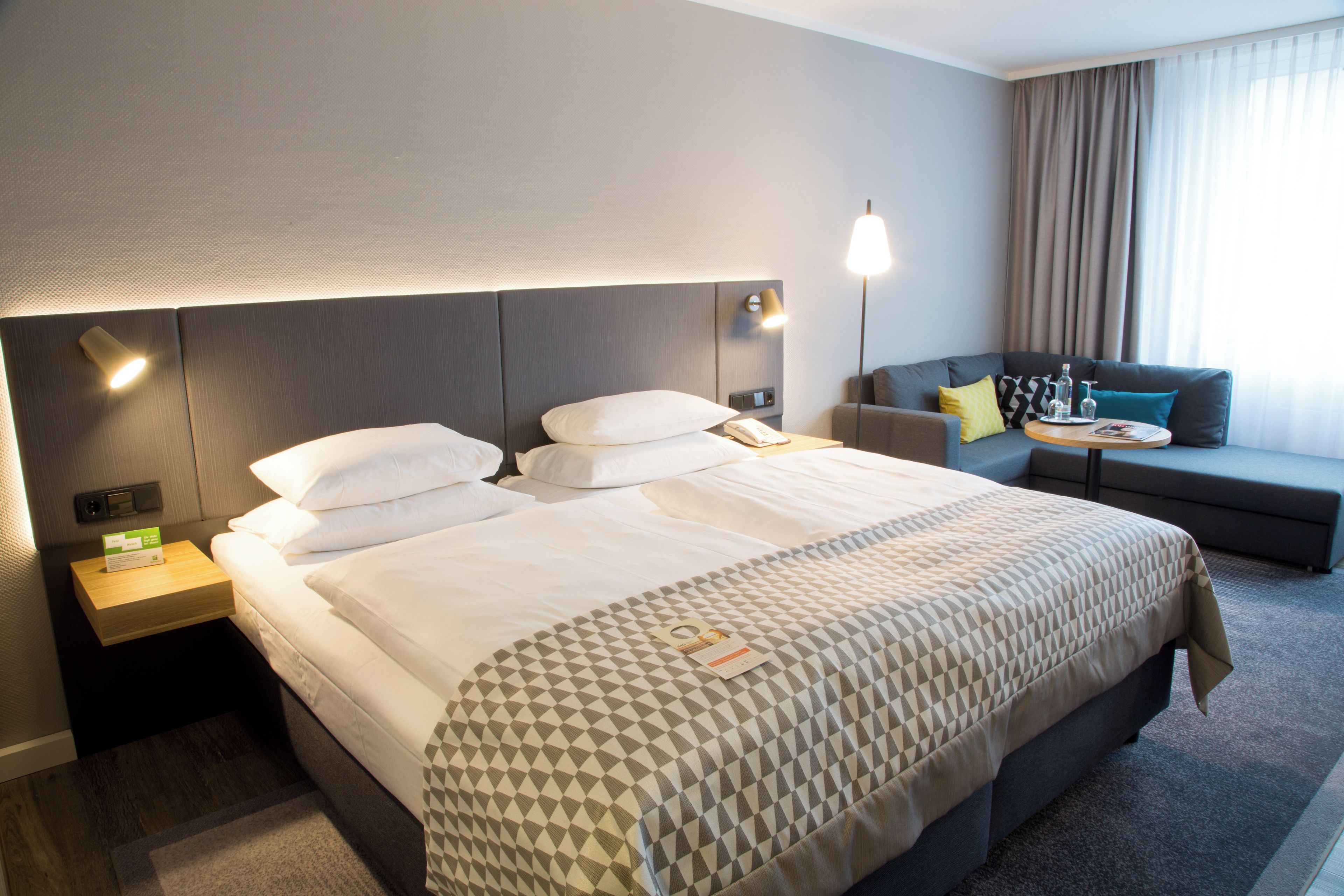 A hotel room with a large bed, a bedside table, a lamp, a sofa, and a window with curtains.