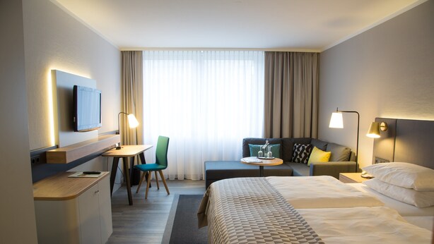 Habitación de hotel con cama, escritorio, sillón, sofá y televisor en la pared.