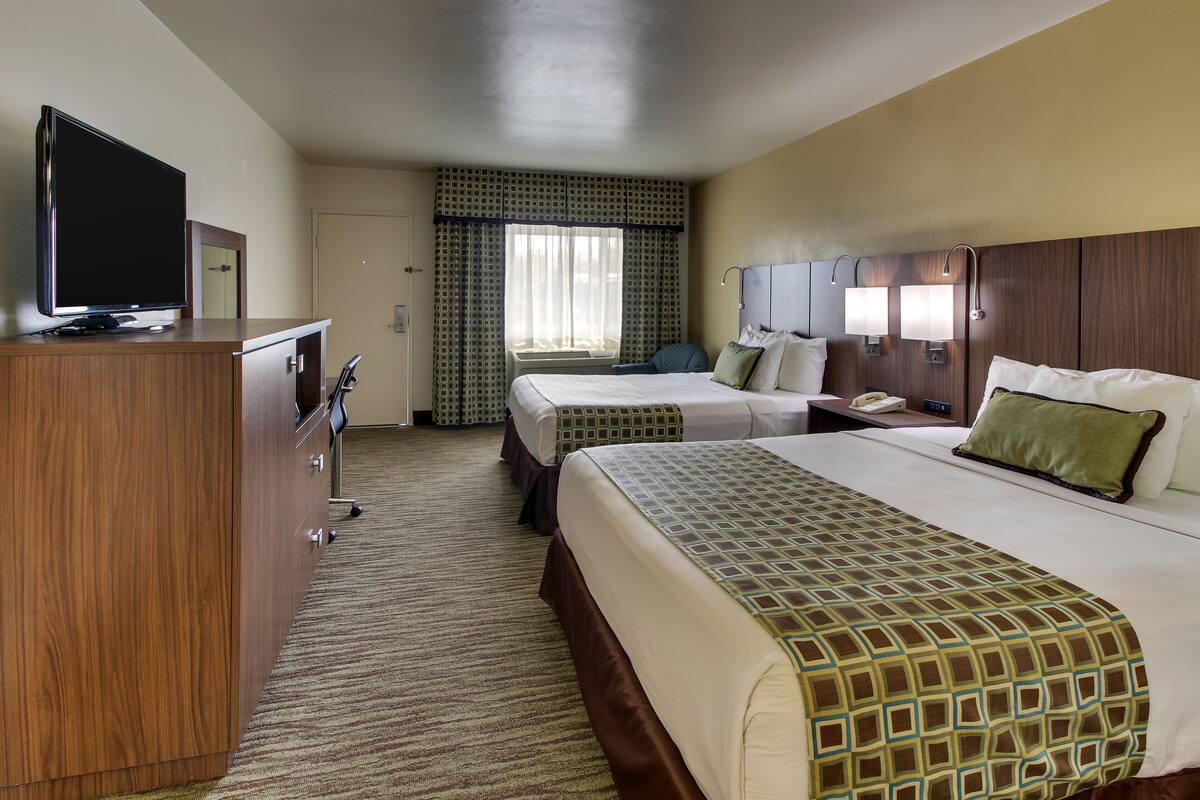 A hotel room with two beds, a TV on a wooden dresser, a desk with a chair, and a window with curtains.