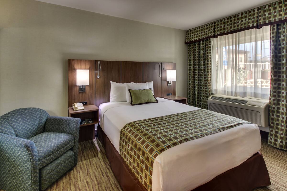 A hotel room with a large bed, a chair, a nightstand, and a window with curtains.