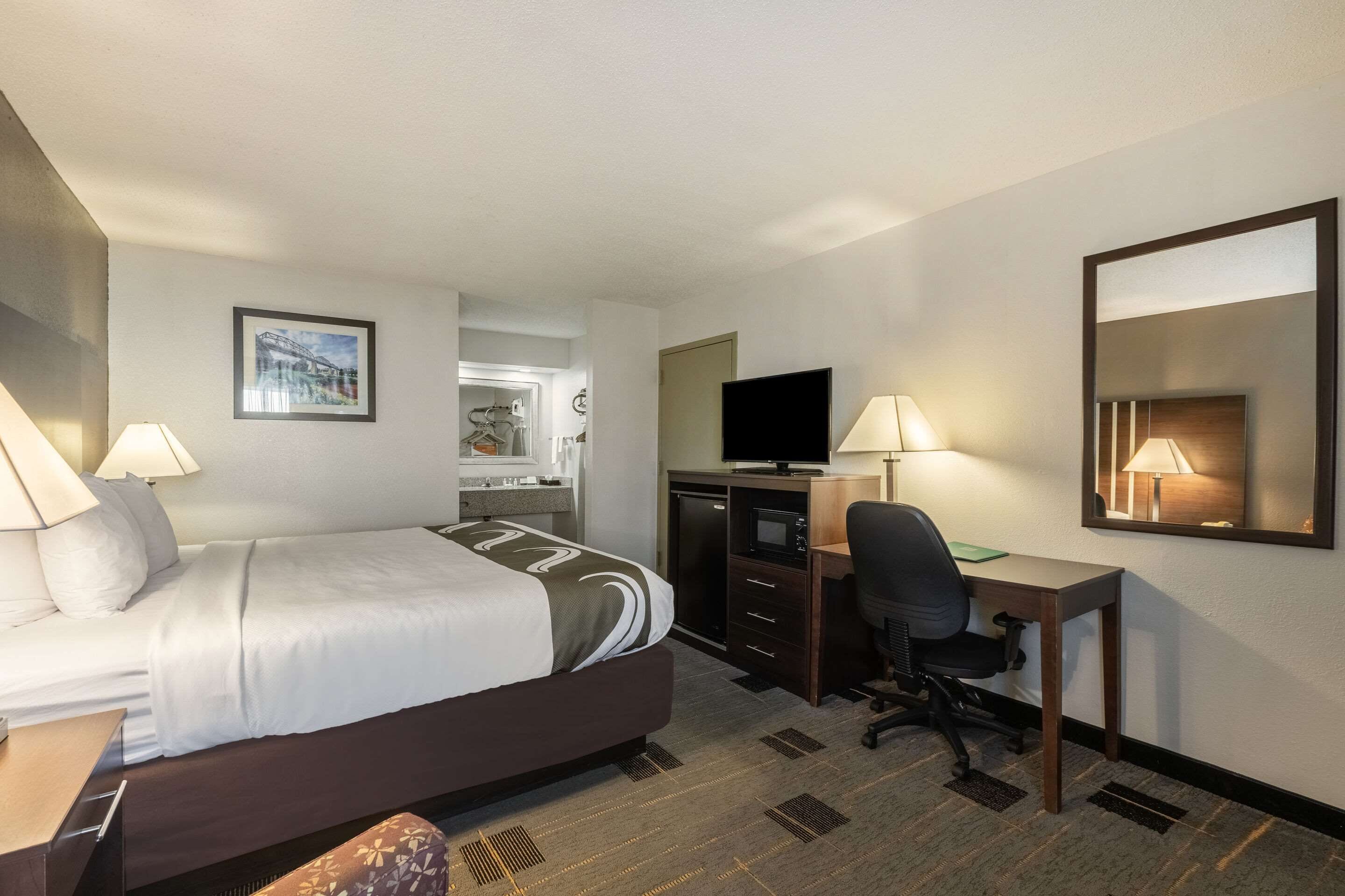 Una habitación de hotel con cama, escritorio, silla, televisor y espejo.