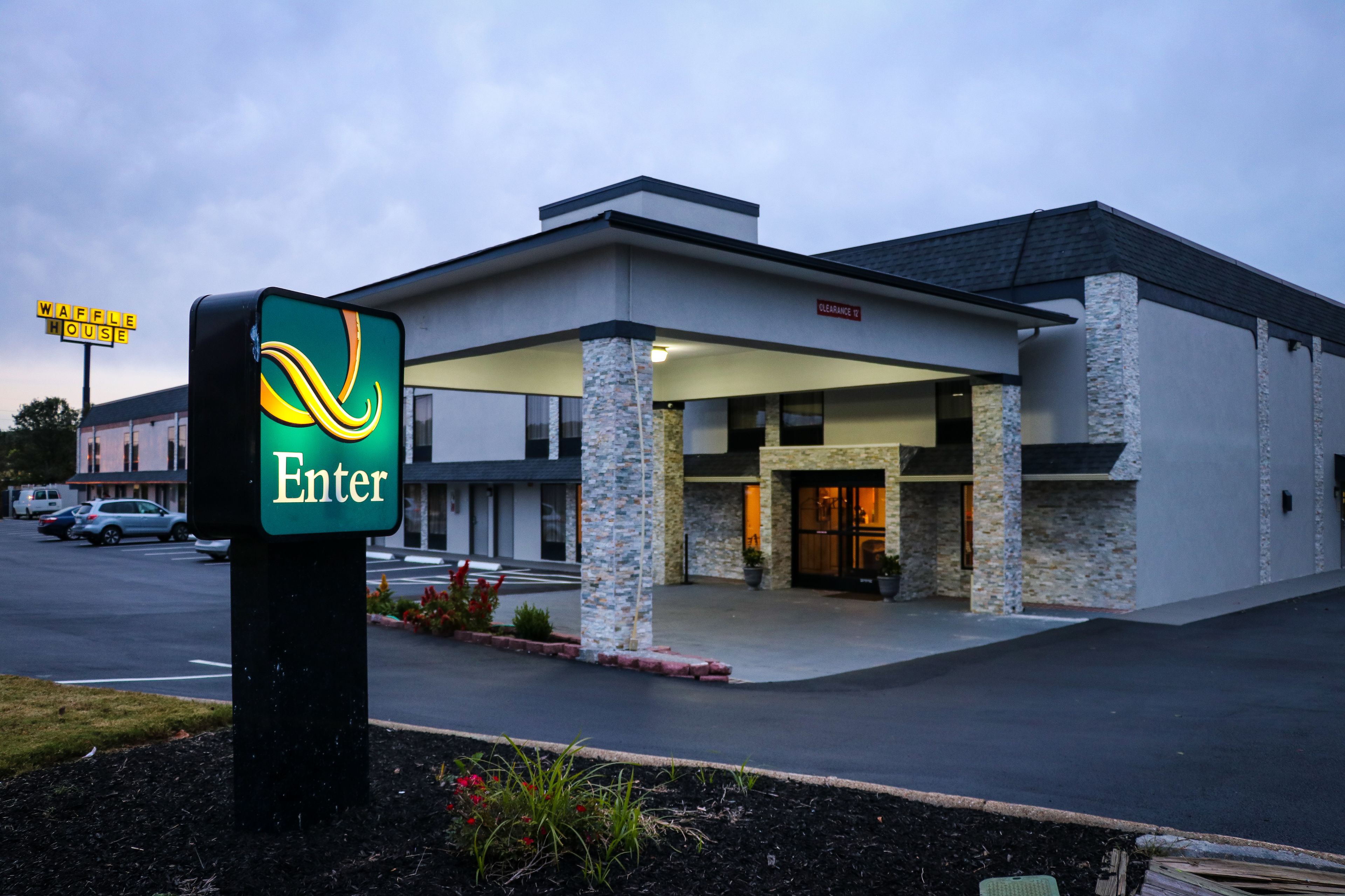 A modern hotel entrance with a clear "Enter" sign, a parking lot, and a visible "Waffle House" sign in the background.