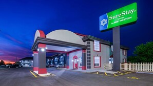 Entrada de hotel con un llamativo letrero SureStay, un área de estacionamiento techada y una cerca blanca de piquetes.