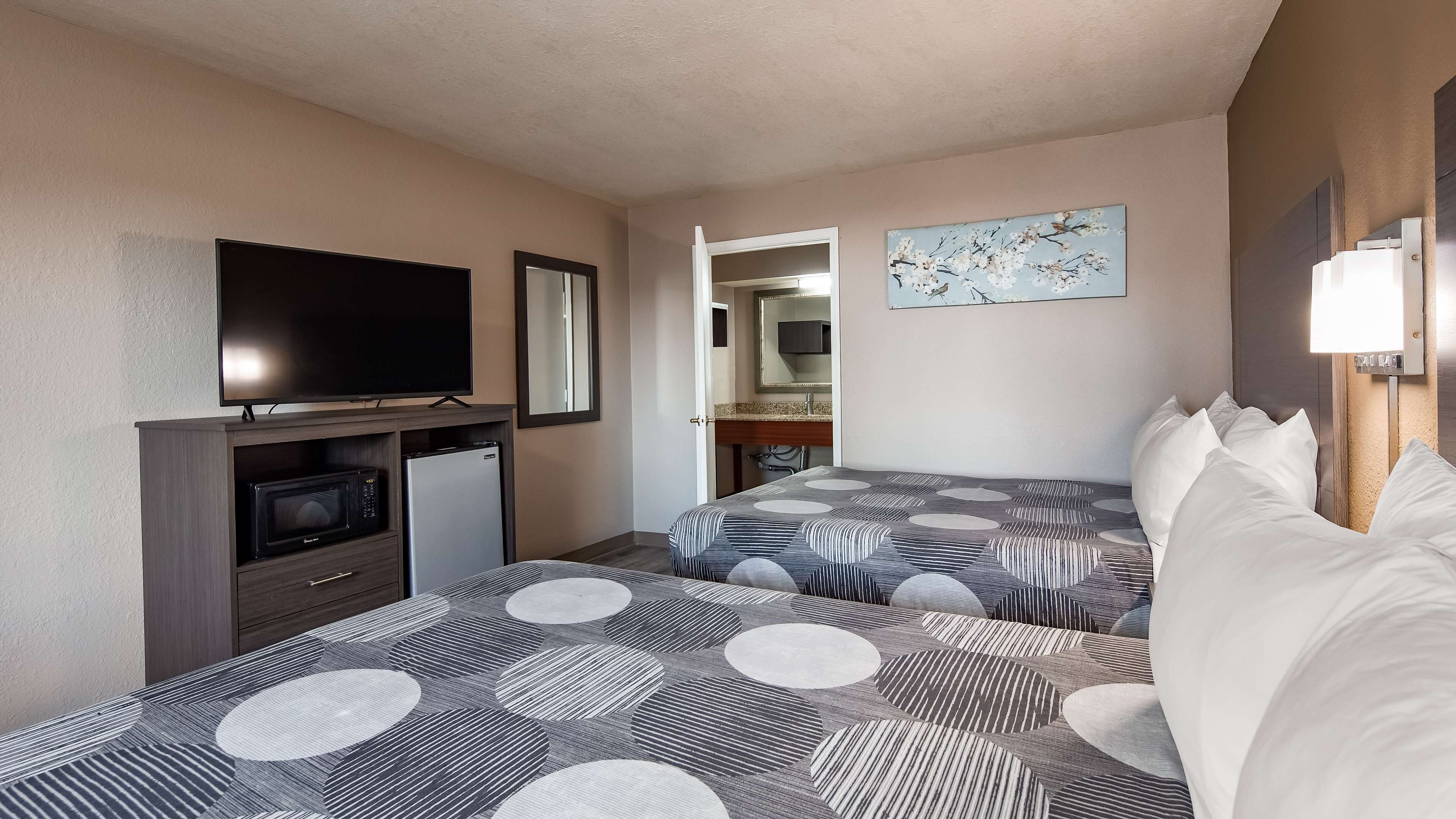 A hotel room with a TV, a small fridge, a bed with patterned bedding, a bedside table, and a painting on the wall.