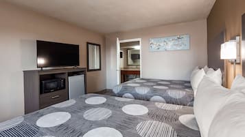 A hotel room with a TV, a small fridge, a bed with patterned bedding, a bedside table, and a painting on the wall.