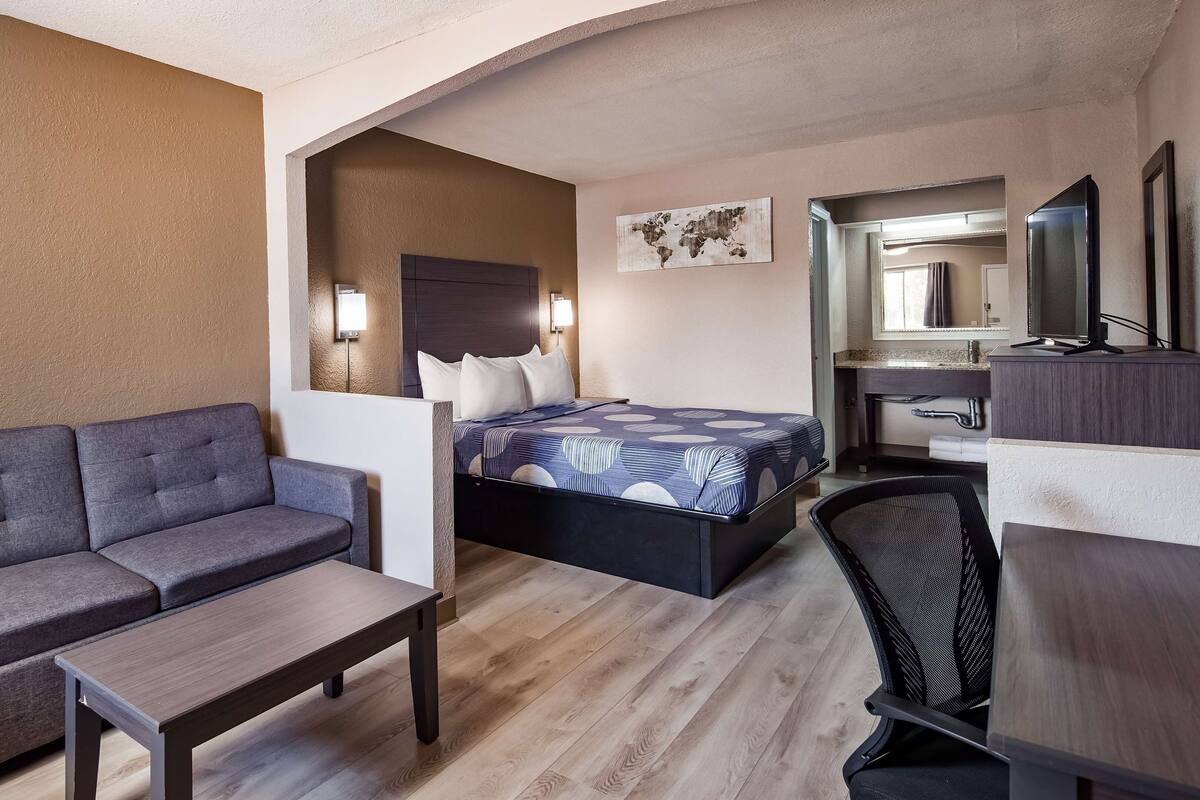 A compact hotel room with a sofa, a bed with a patterned bedspread, a desk with a chair, and a mirror.