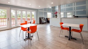 Amplio espacio interior con ventanas grandes, un mostrador de recepción y dos mesas con sillas naranjas.