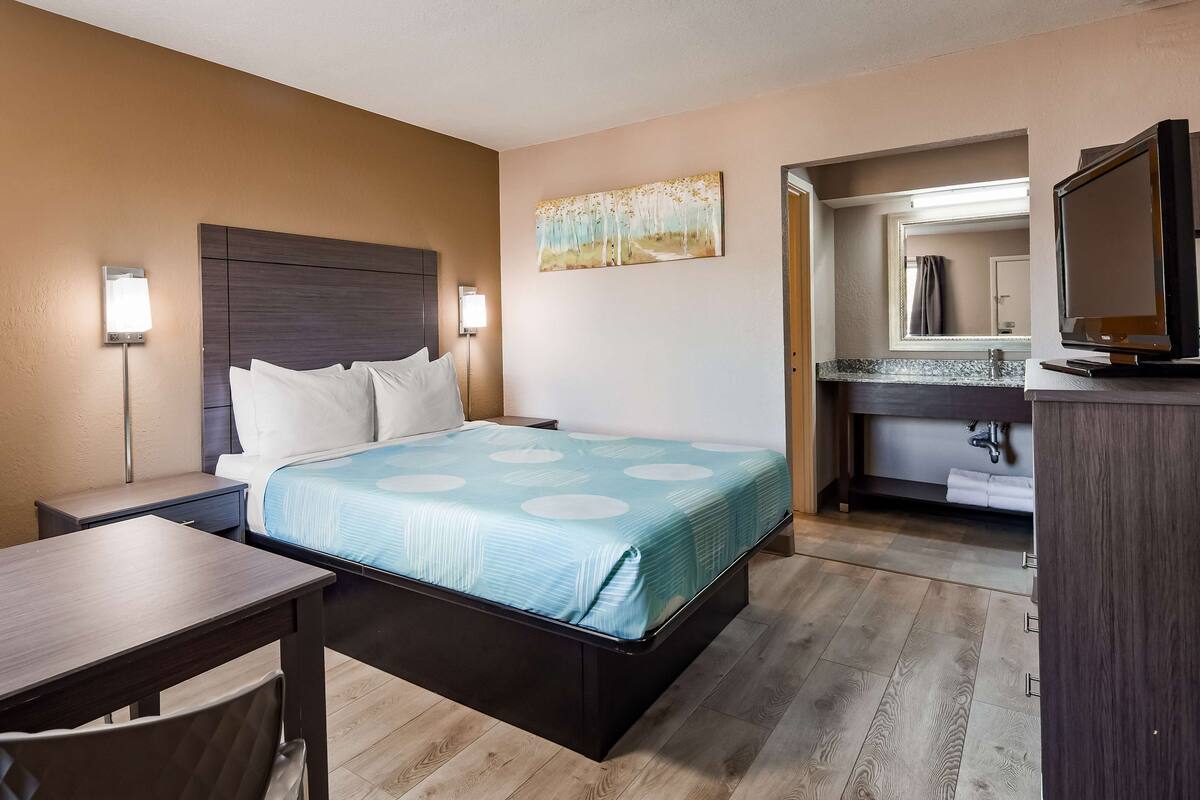A hotel room with a bed, a desk, a chair, a nightstand, a wall-mounted TV, and a bathroom visible through an open door.