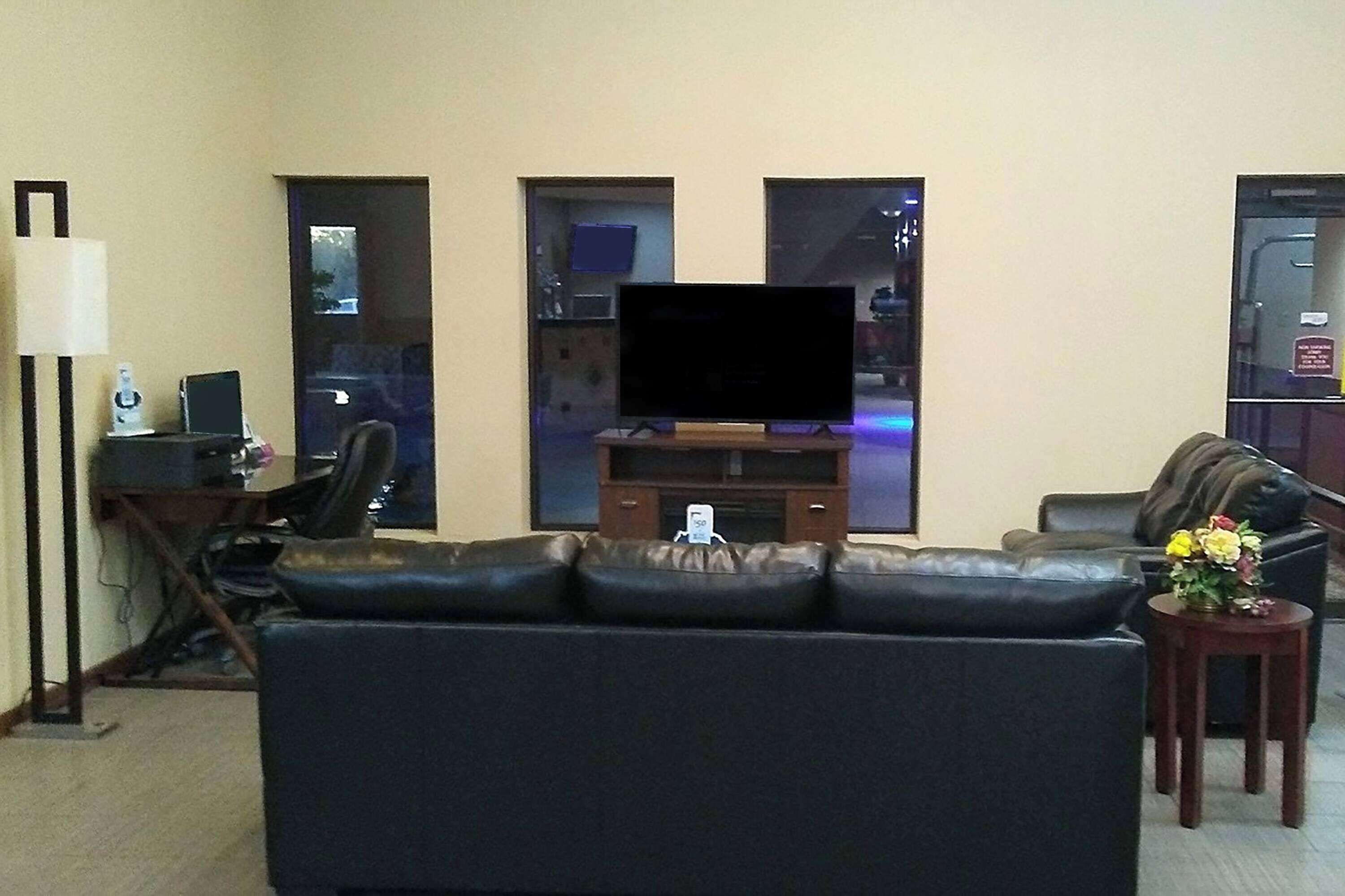 Una sala de estar con un sofá de cuero negro, un televisor de pantalla plana sobre un soporte de madera, una mesita con un jarrón de flores y un escritorio con una computadora y una lámpara.