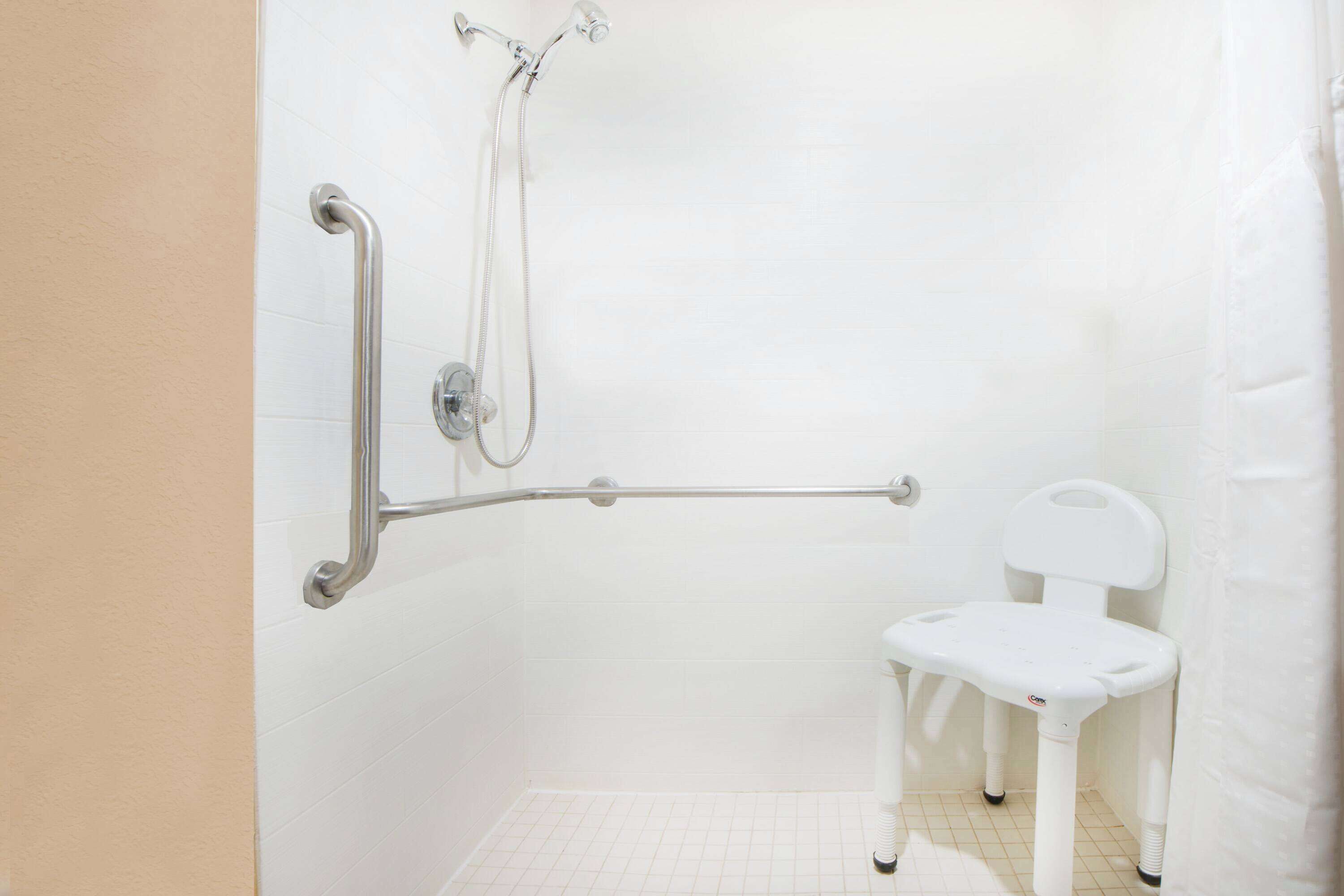 Un baño con pared de azulejos blancos, una ducha con cabezal de ducha manual, una barra de apoyo y una silla de plástico blanca.