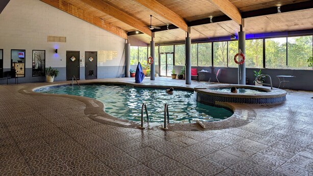 Una piscina interior con vigas de madera, suelo de baldosas y vistas a árboles.