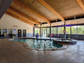 An indoor swimming pool with wooden beams, tiled flooring, and a view of trees outside.