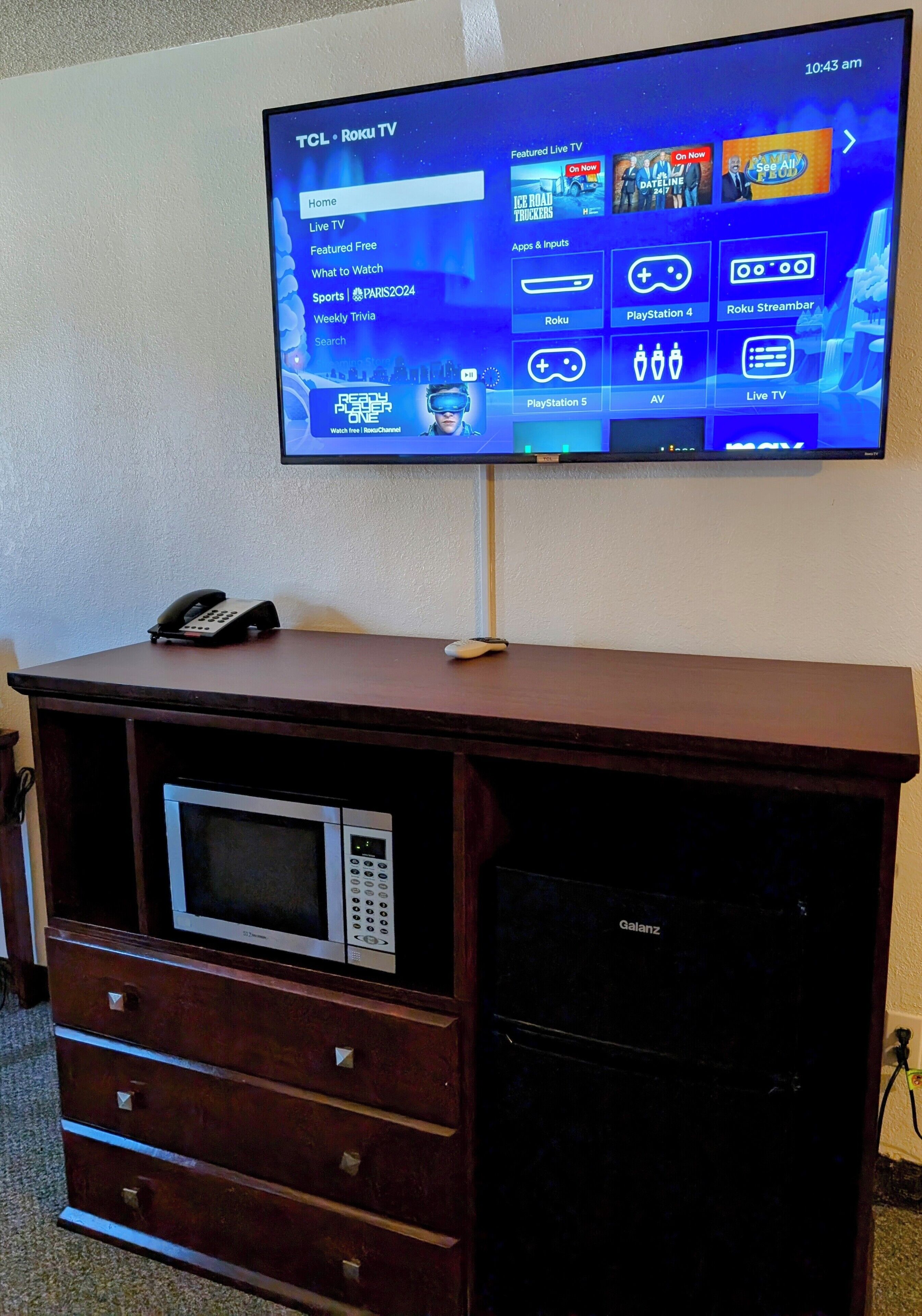 Ein TCL Roku Fernseher an einem Holzschrank, darunter eine Mikrowelle und Schubladen.