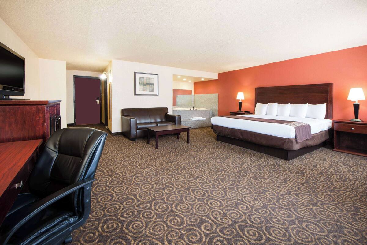 A hotel room with a bed, a desk, a chair, a TV, and a bathroom visible through an open door.