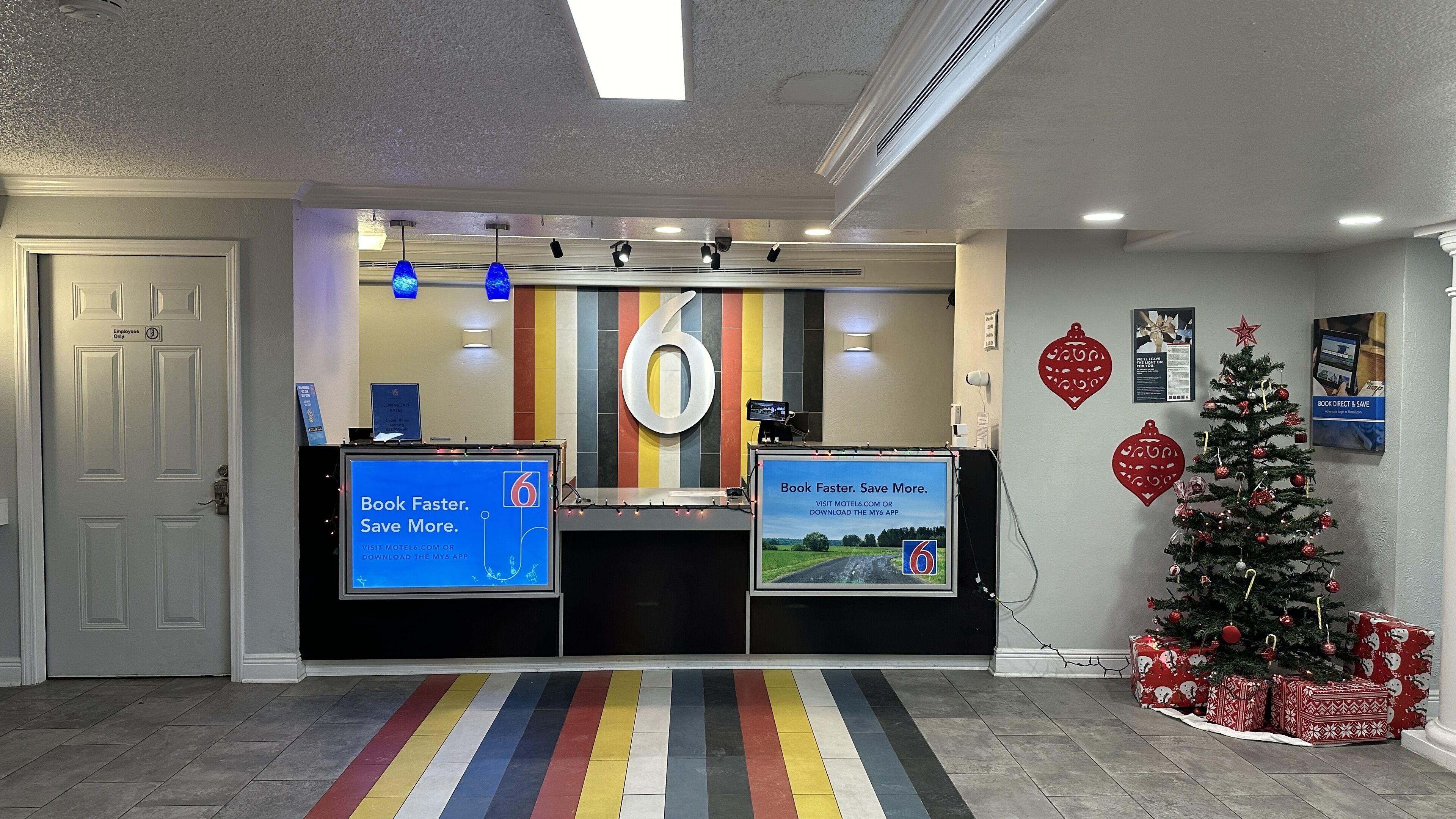 Un hall d’accueil d’hôtel avec un grand panneau portant le chiffre « 6 », un sapin de Noël et un tapis rayé aux couleurs vives.