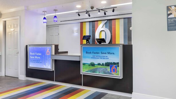 A hotel reception area with a digital screen displaying a booking message, a large number '6' on the wall, and a colorful striped carpet. - Motel 6 Beaumont, TX (Beaumont)