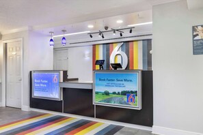 A hotel reception area with a digital screen displaying a booking message, a large number '6' on the wall, and a colorful striped carpet. - Motel 6 Beaumont, TX (Beaumont)