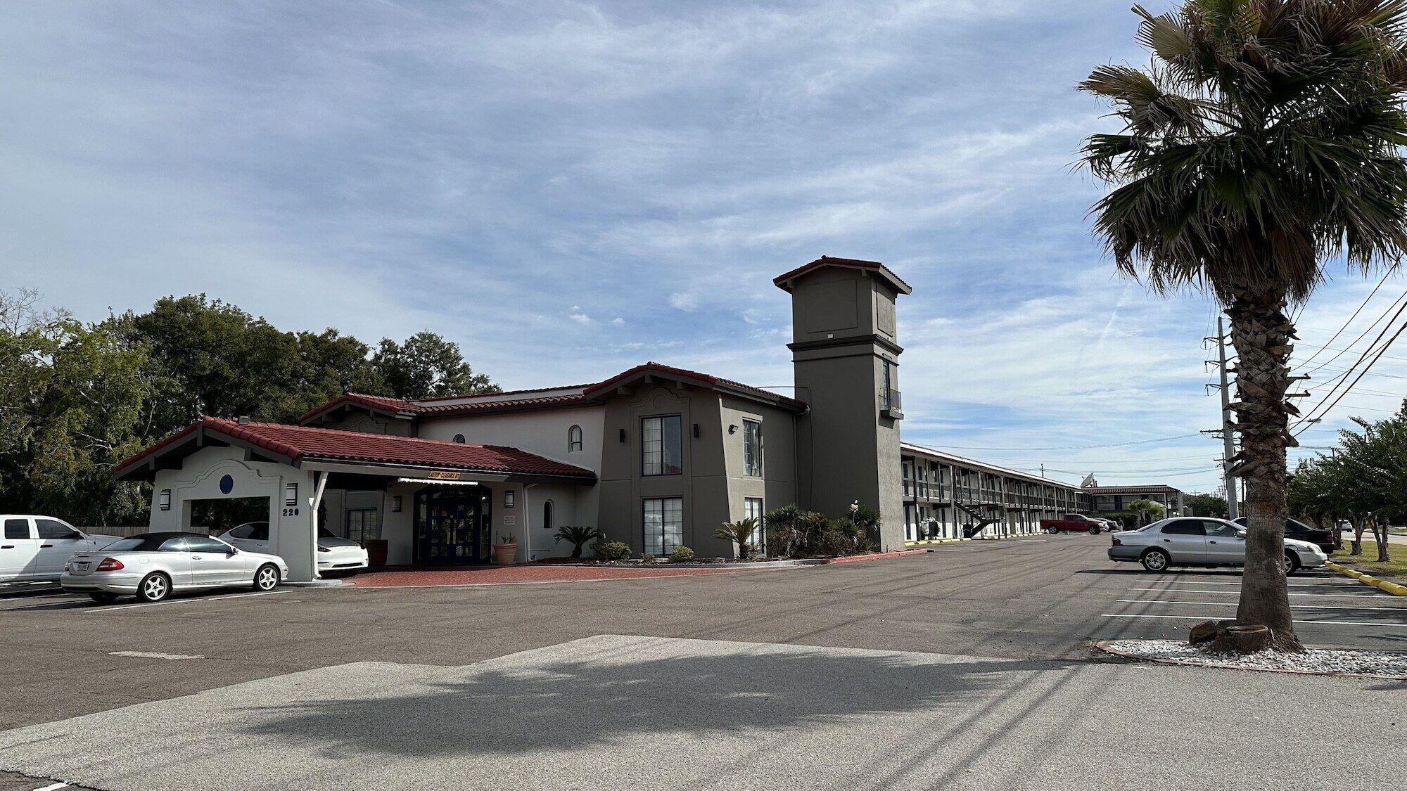 A two-story building with a red-tiled roof and a tall tower. There are cars parked in front and a palm tree to the right.