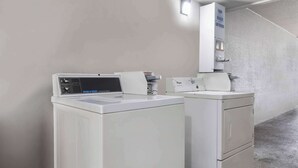 A laundry area with a commercial washer and dryer, a soap and bleach softener dispenser, and a wall-mounted light fixture. - Motel 6 Beaumont, TX (Beaumont)