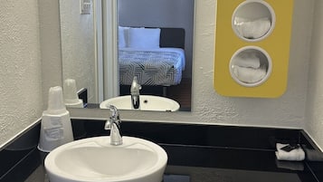 Un baño con lavamanos blanco, encimera negra y un espejo. Hay un perchero con tres toallas y una botella de jabón de manos sobre la encimera.