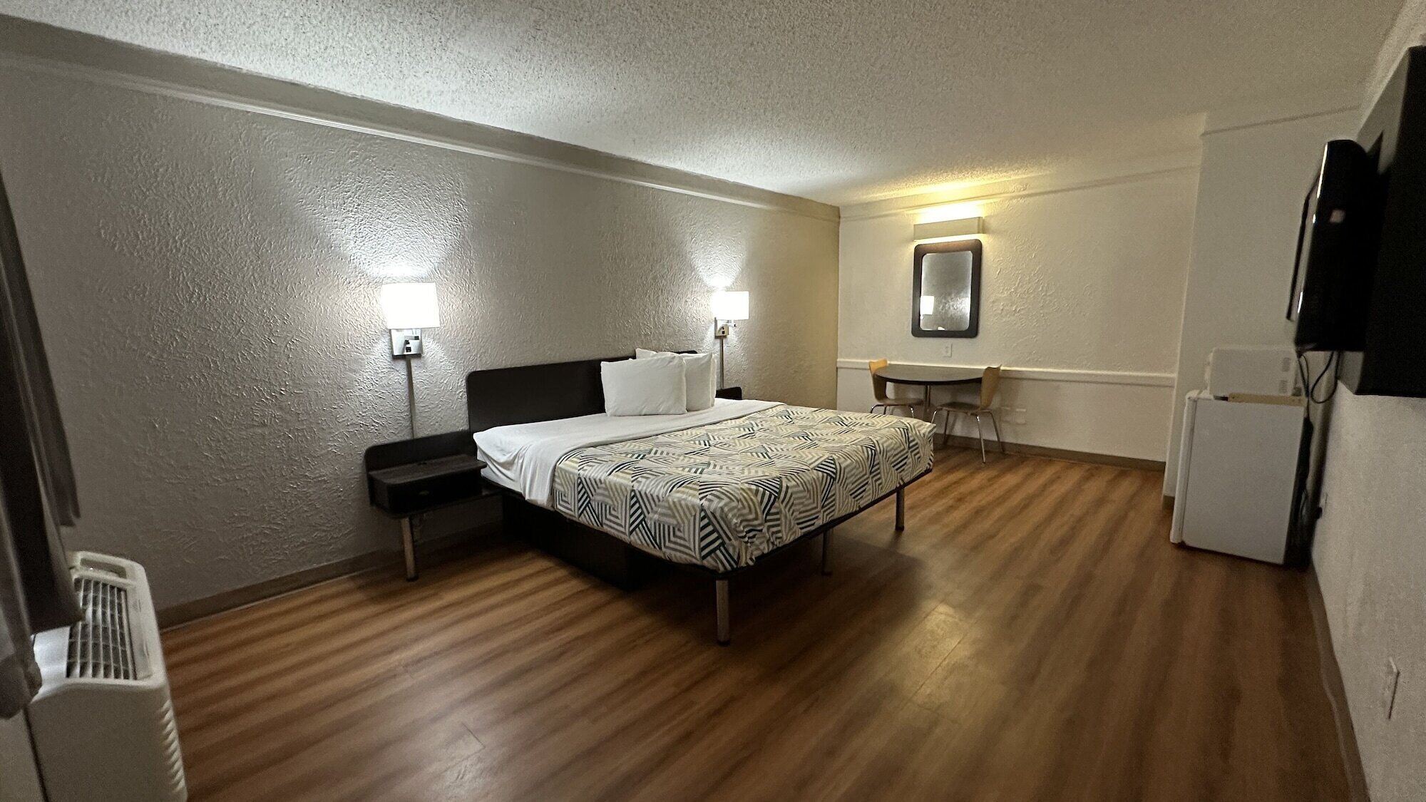 A hotel room with a bed, bedside table, lamp, small desk, chair, and a refrigerator.