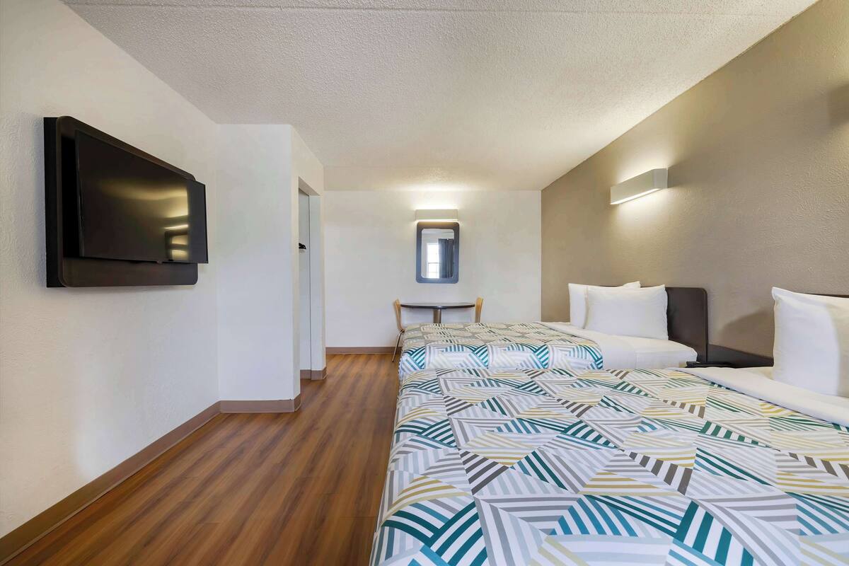 A hotel room with a bed, a flat-screen TV mounted on the wall, a small table, and a window with a mirror above it.