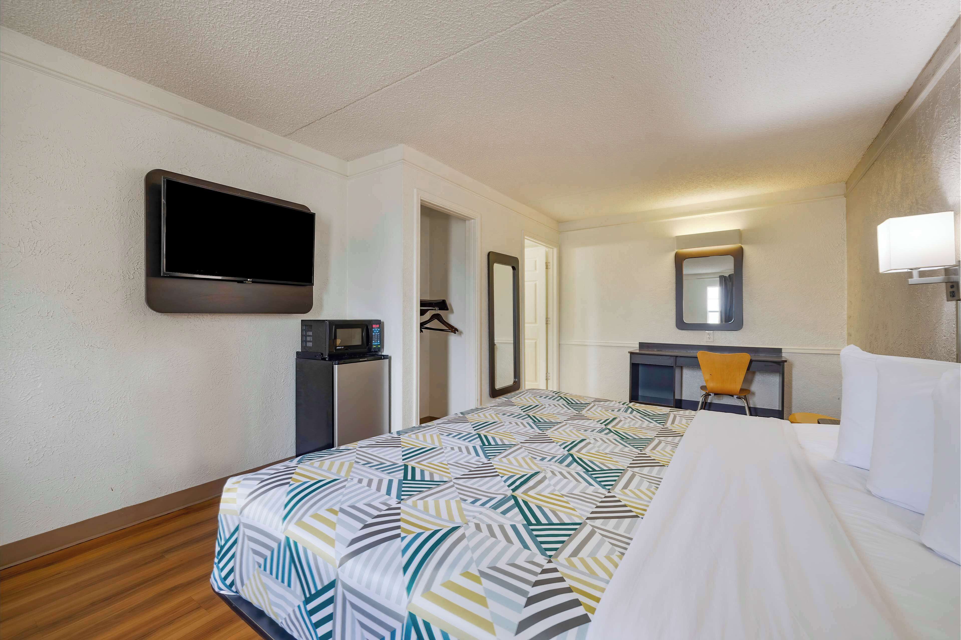 A hotel room with a bed, a TV mounted on the wall, a desk with a chair, a microwave, and a mirror.