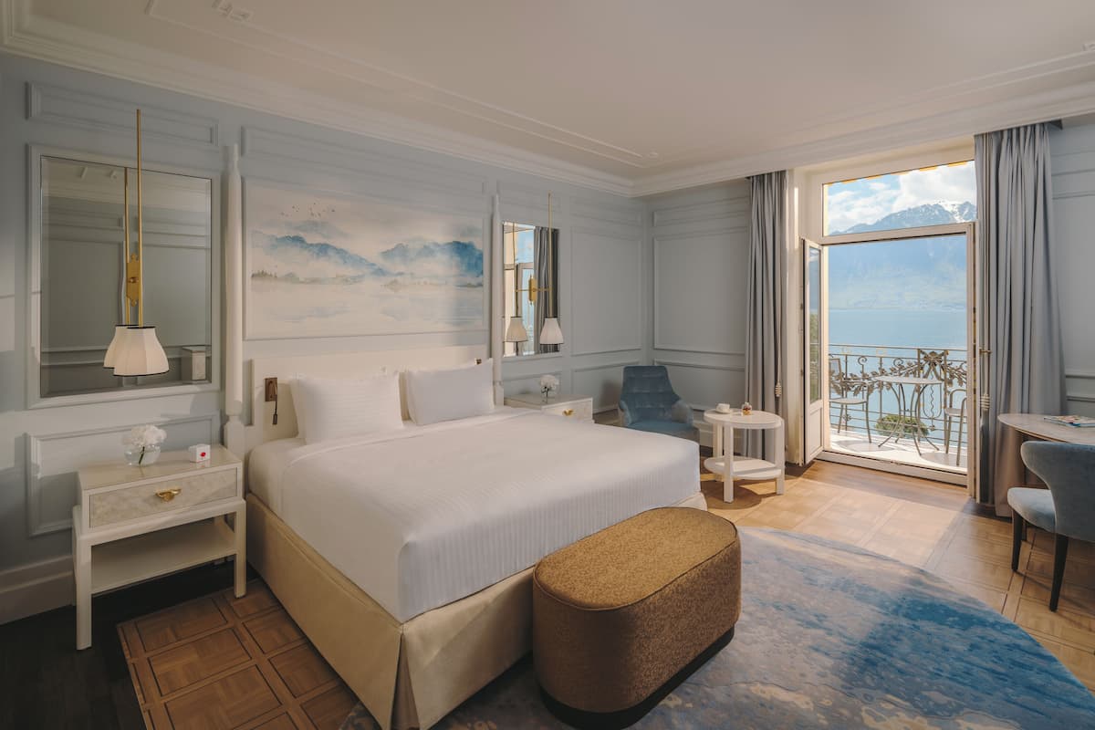 A hotel room with a large bed, a nightstand, a chair, a small table, and a view of the sea through a balcony.