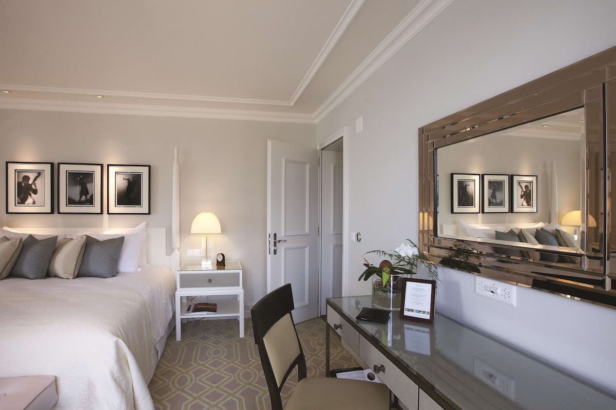 A hotel room with a bed, bedside table, lamp, chair, desk, mirror, and framed pictures on the wall.