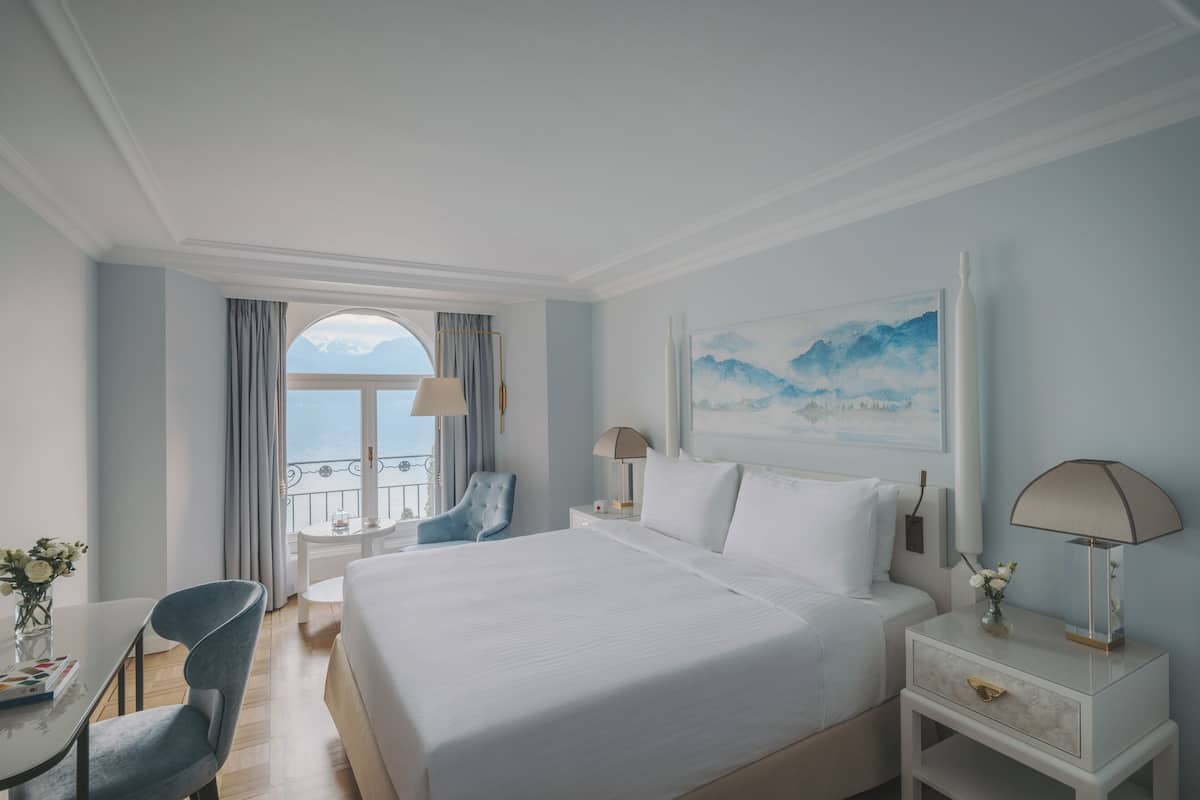 Una habitación de hotel con una cama grande, un escritorio con silla, una mesita de noche con lámpara y una ventana con vistas al mar.