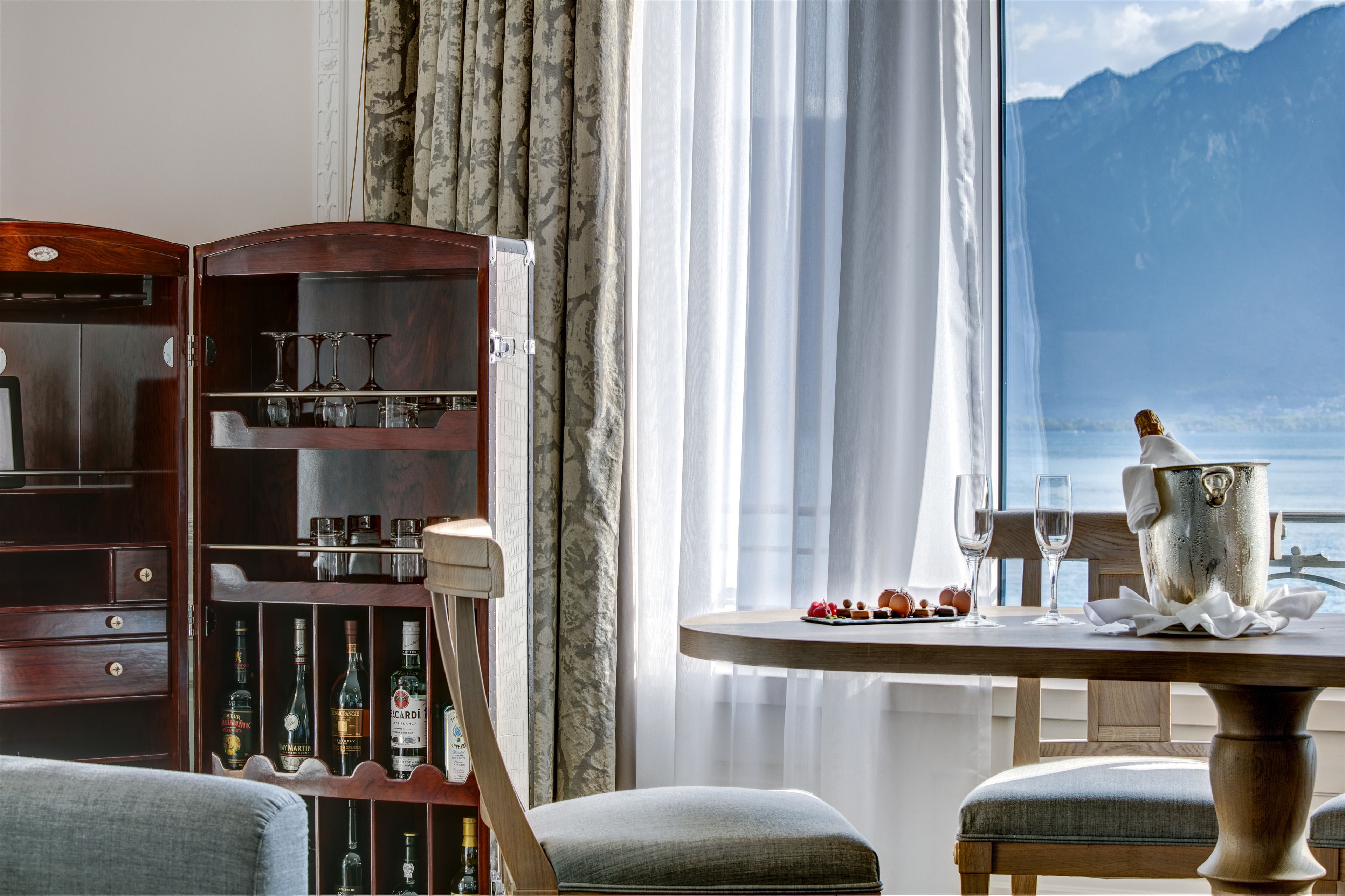 A room with a wooden cabinet containing bottles, a table set for two with glasses and a silver pot, and a view of mountains through a window.