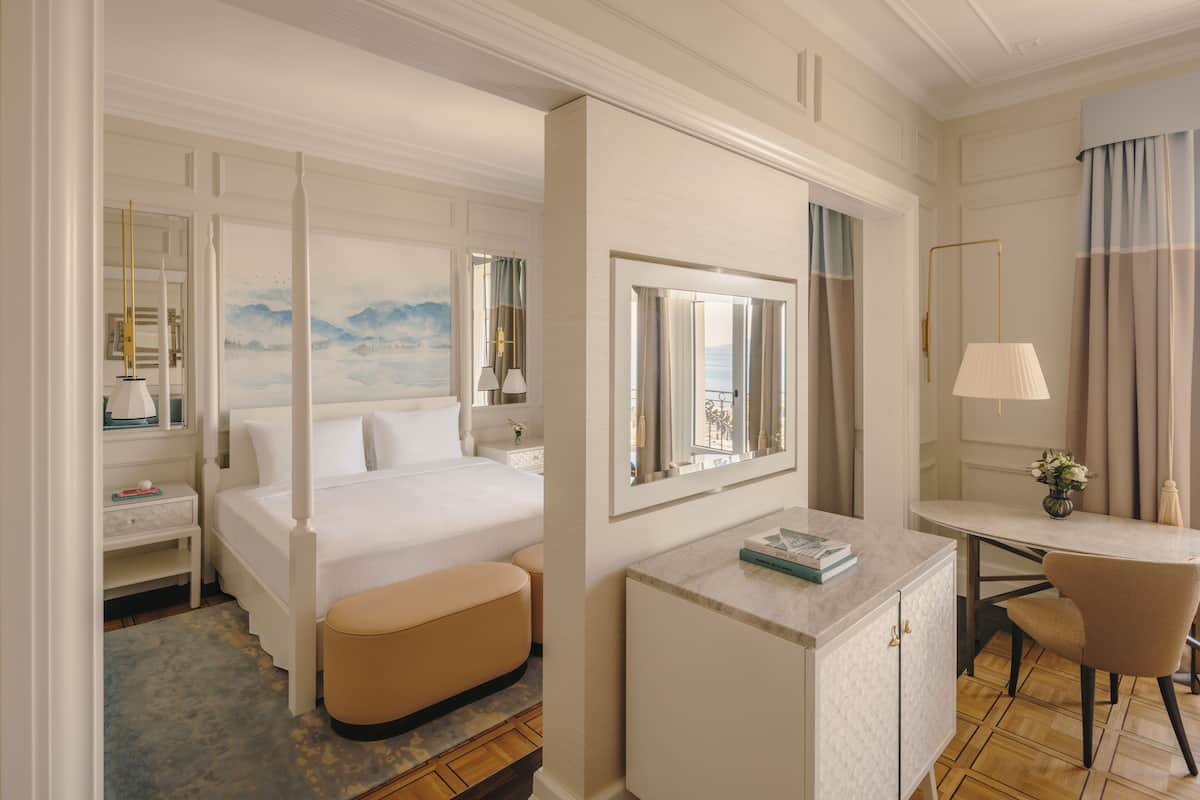 A hotel room with a four-poster bed, a sitting area with a cushioned bench, a desk with a chair, and a large mirror above the desk.