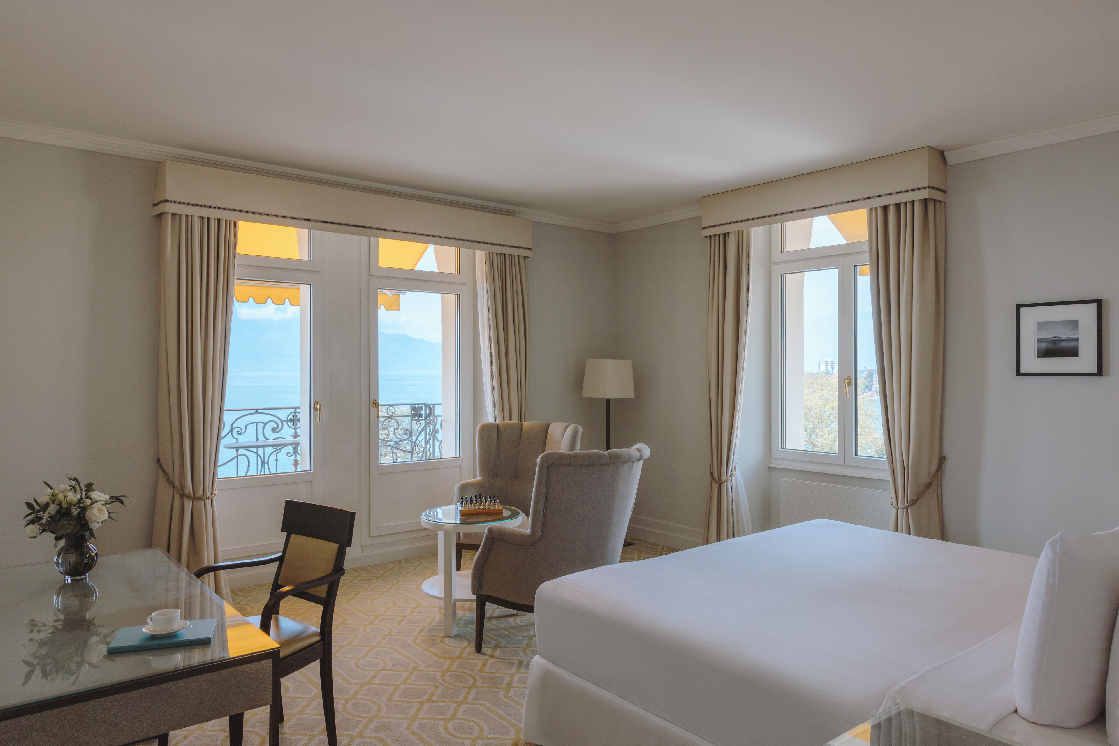 A hotel room with a bed, a chair, a small table with a vase of flowers, and a view of the sea through large windows.
