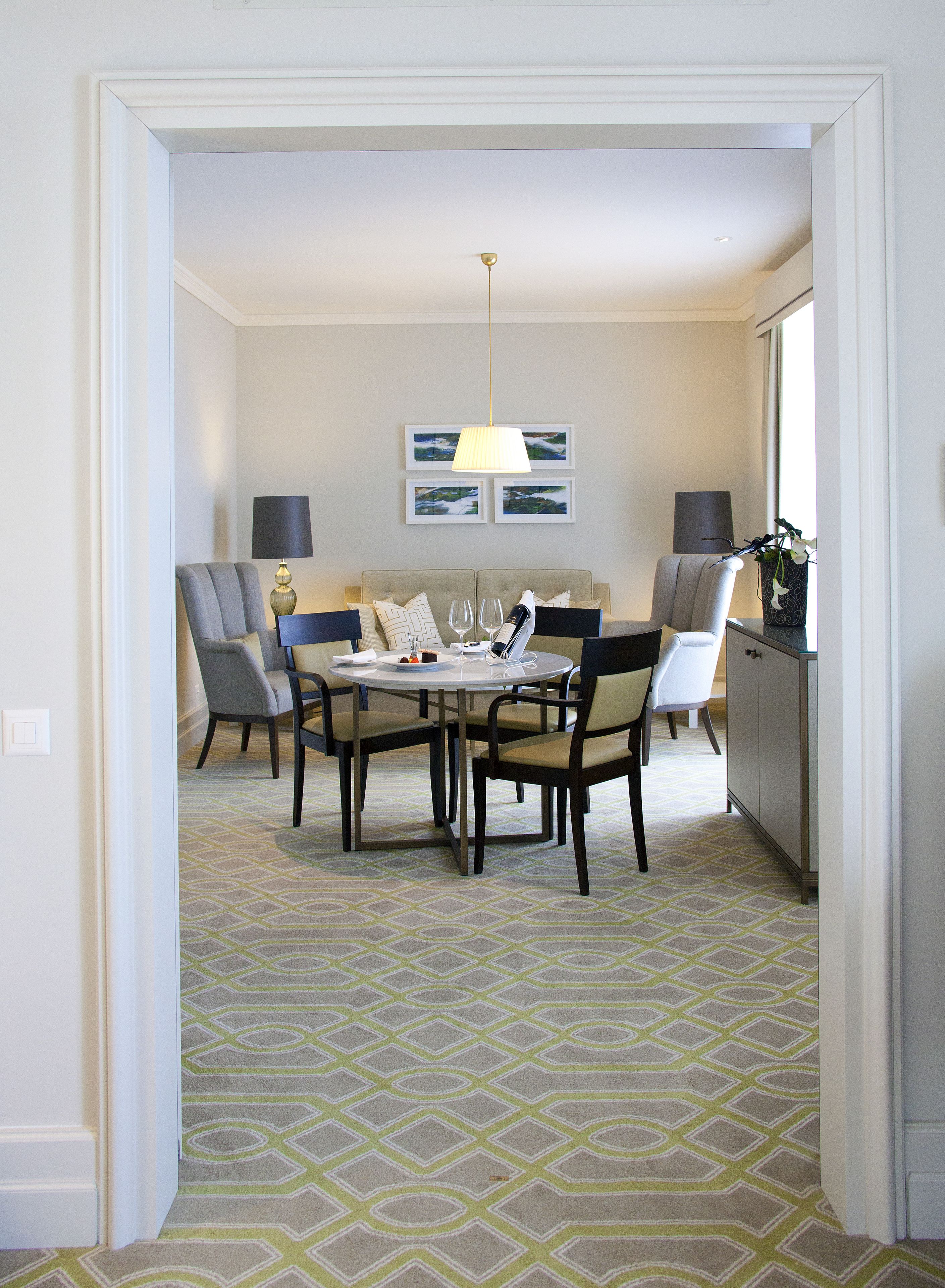 A dining area with a round table, chairs, and a chandelier. There are framed pictures on the wall and a cabinet with a plant.