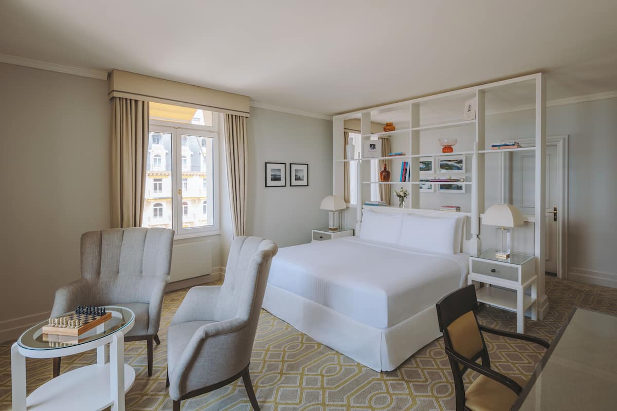Habitación de hotel con una cama grande, dos sillones, una mesita con ajedrez, una mesita de noche con lámparas y un armario con estantes.